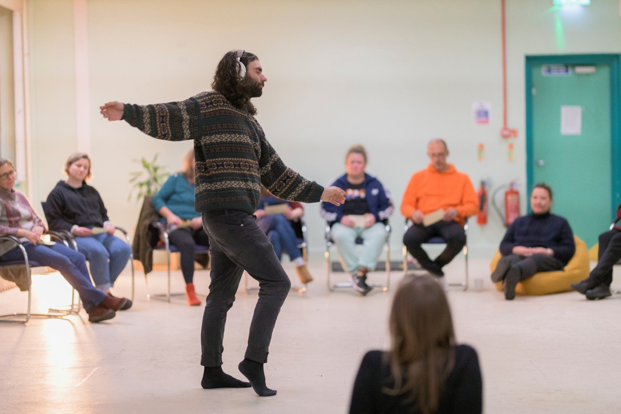 A group of people sitting in a circle on chairs with a peson dancing in the middle of the circle wearing headphones as part of the And Then residency