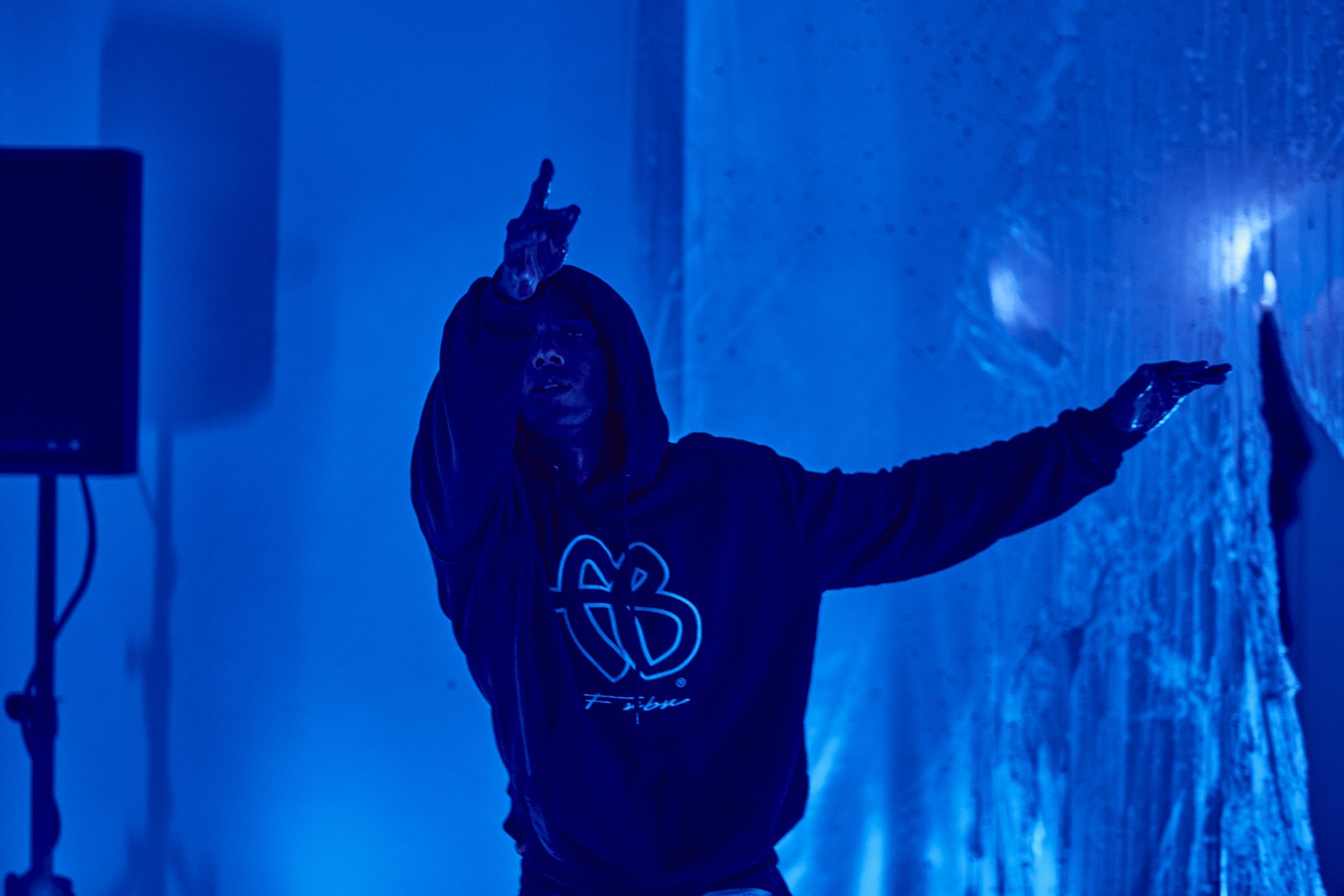 A photo of Malik Nashad-Sharpe dancing, wearing a hoodie in a room lit in blue