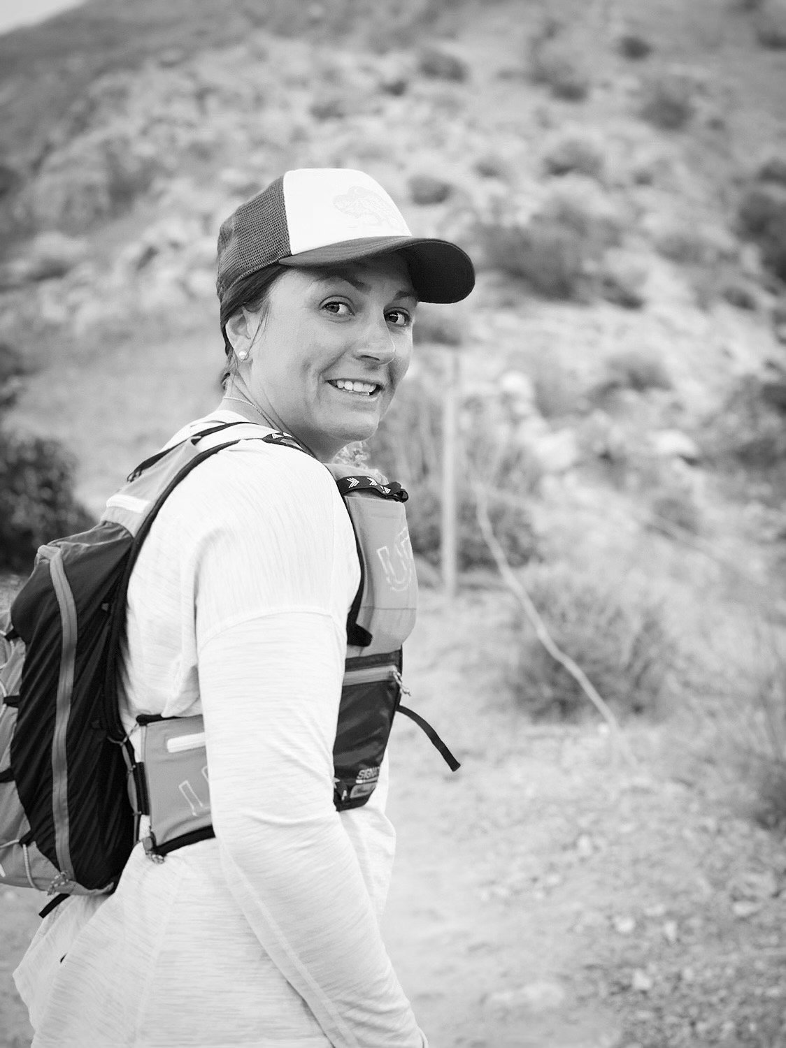 A woman in outdoor hiking gear, smiling, with a backpack, standing on a trail with a rocky hillside background.