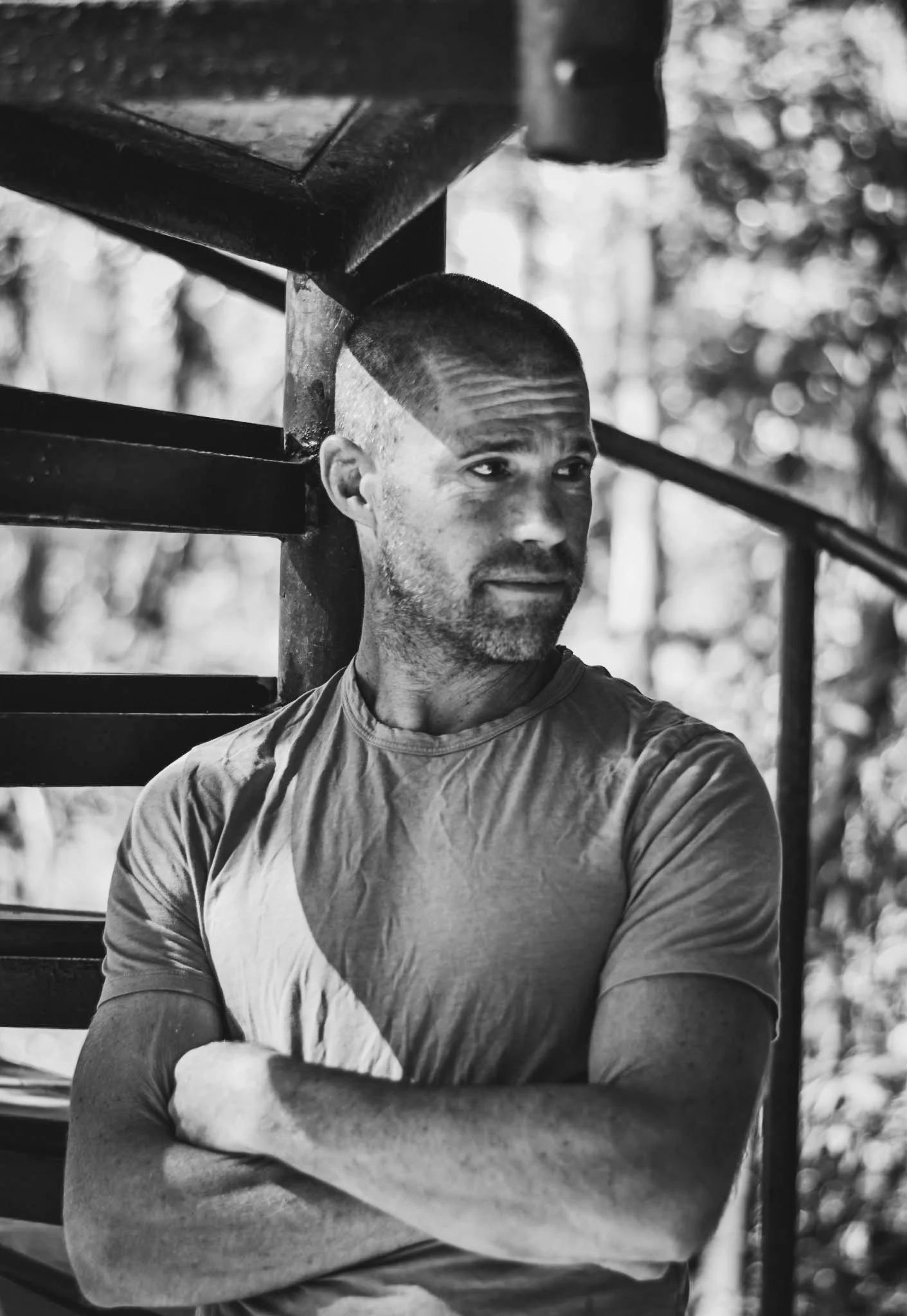 A man with a shaved head and beard standing with his arms crossed outdoors, looking to the side, with trees in the background.