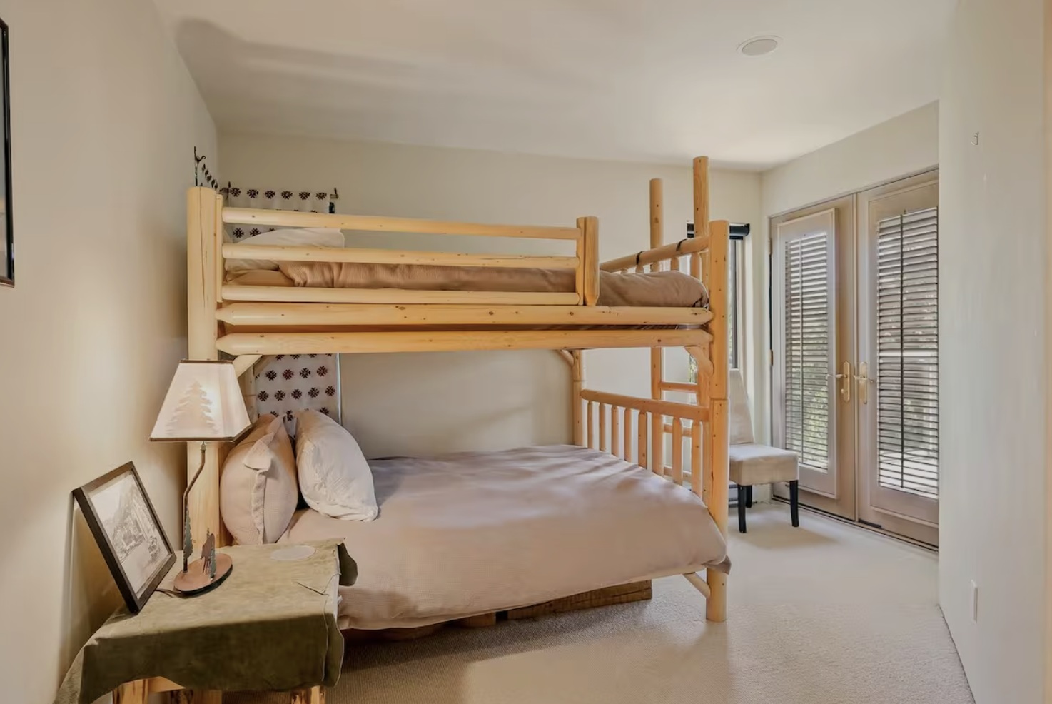 Bedroom with a wooden bunk bed, a nightstand with a lamp and framed picture, and a chair near glass doors with shutters.
