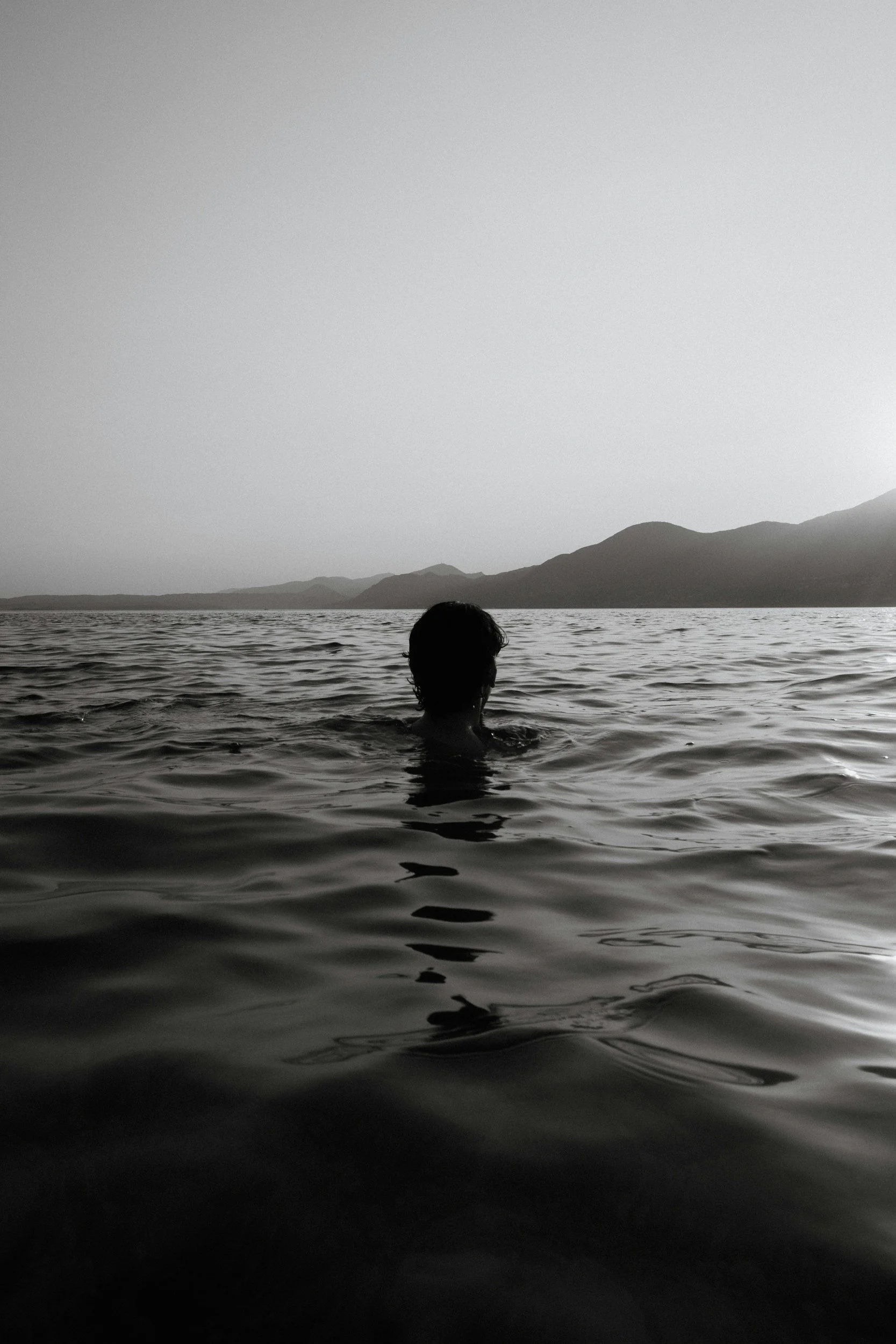 A person with short curly hair swimming in a calm body of water with distant mountains in the background, black and white photograph.