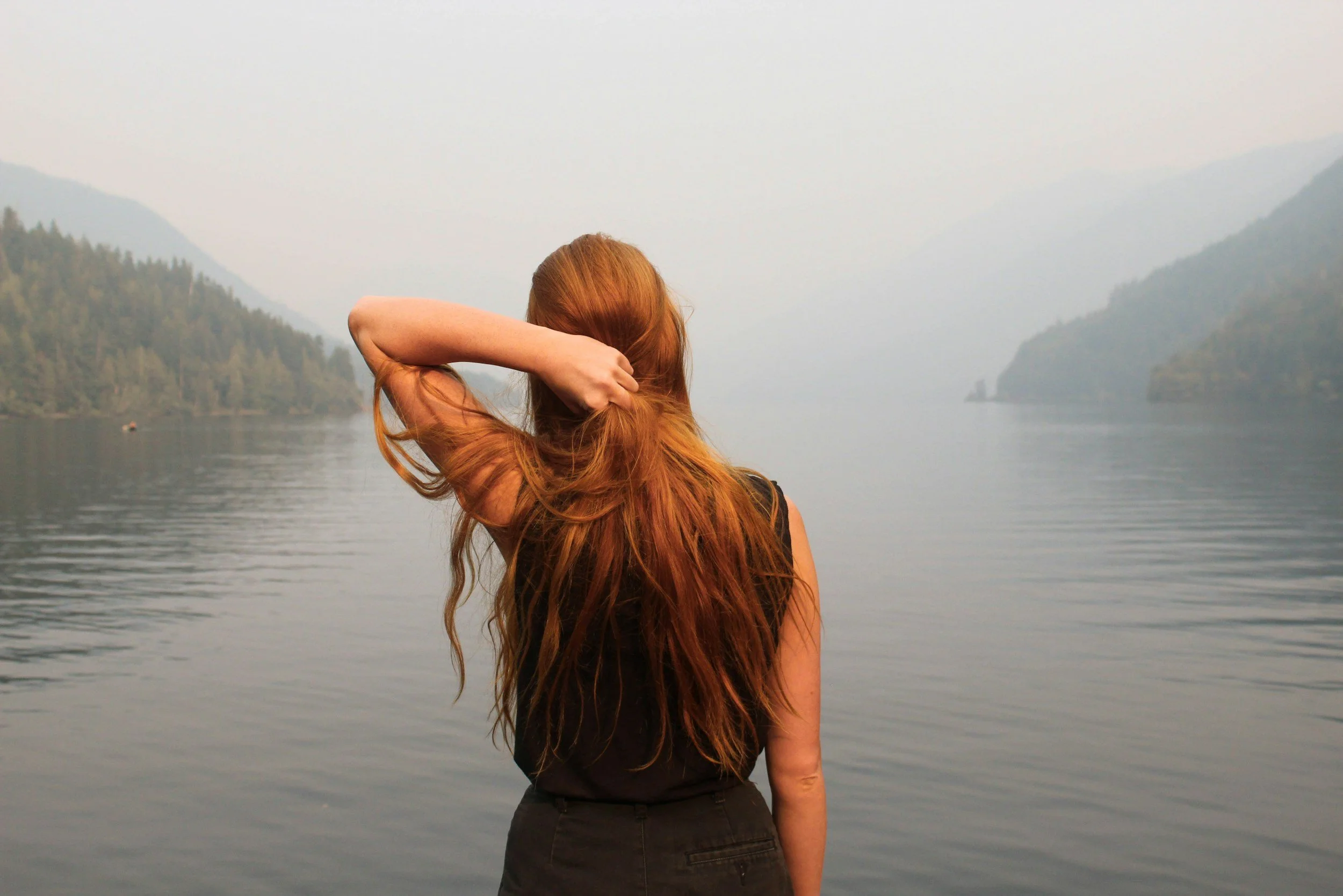 A woman with long red hair, wearing a black top and black pants, facing away and standing near a body of water with mountains in the background.