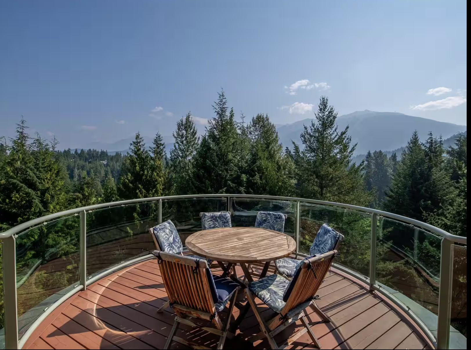 Outdoor wooden deck with a round table and six chairs with floral cushions, overlooking a forested mountain landscape with trees and distant mountains under a clear blue sky.
