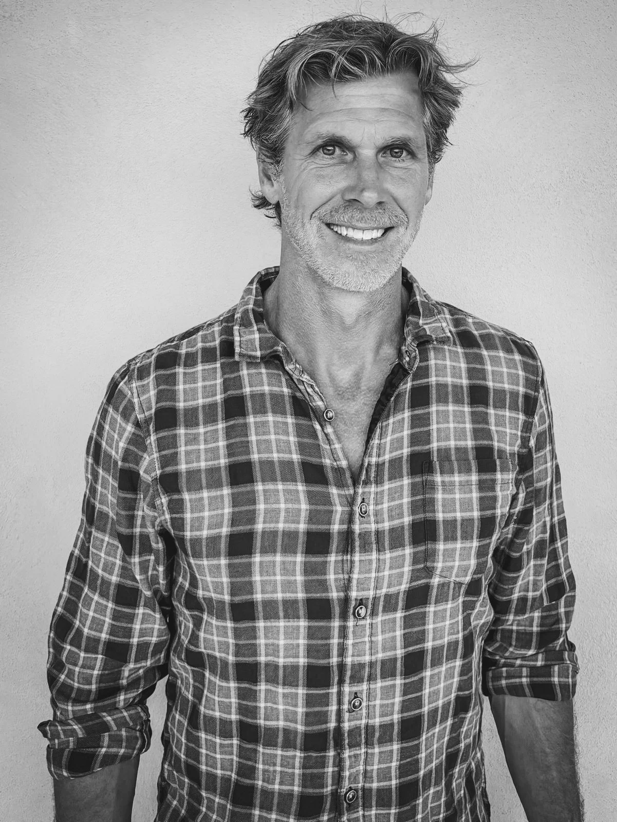 Black and white photo of a smiling man with wavy hair and a beard, wearing a plaid button-up shirt, standing against a plain wall.