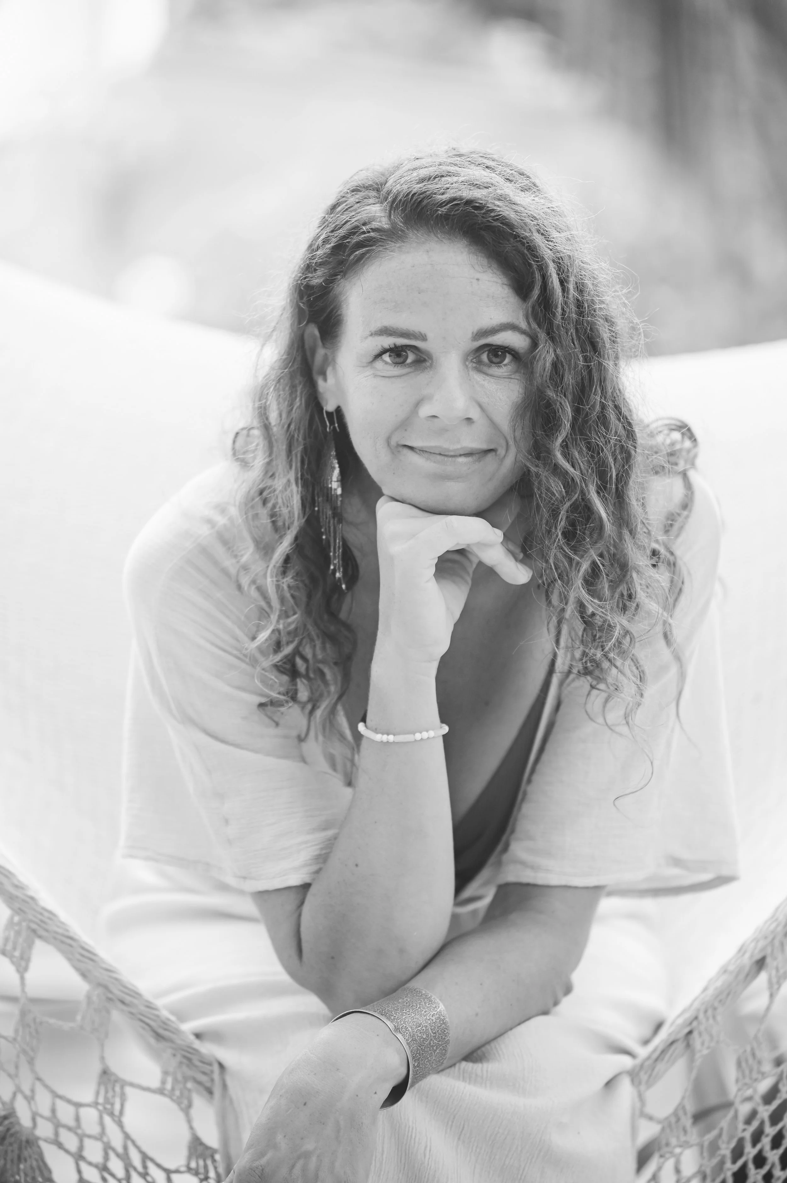 Black and white photo of a woman with curly hair, resting her chin on her hand, looking at the camera with a slight smile, wearing earrings and a bracelet.