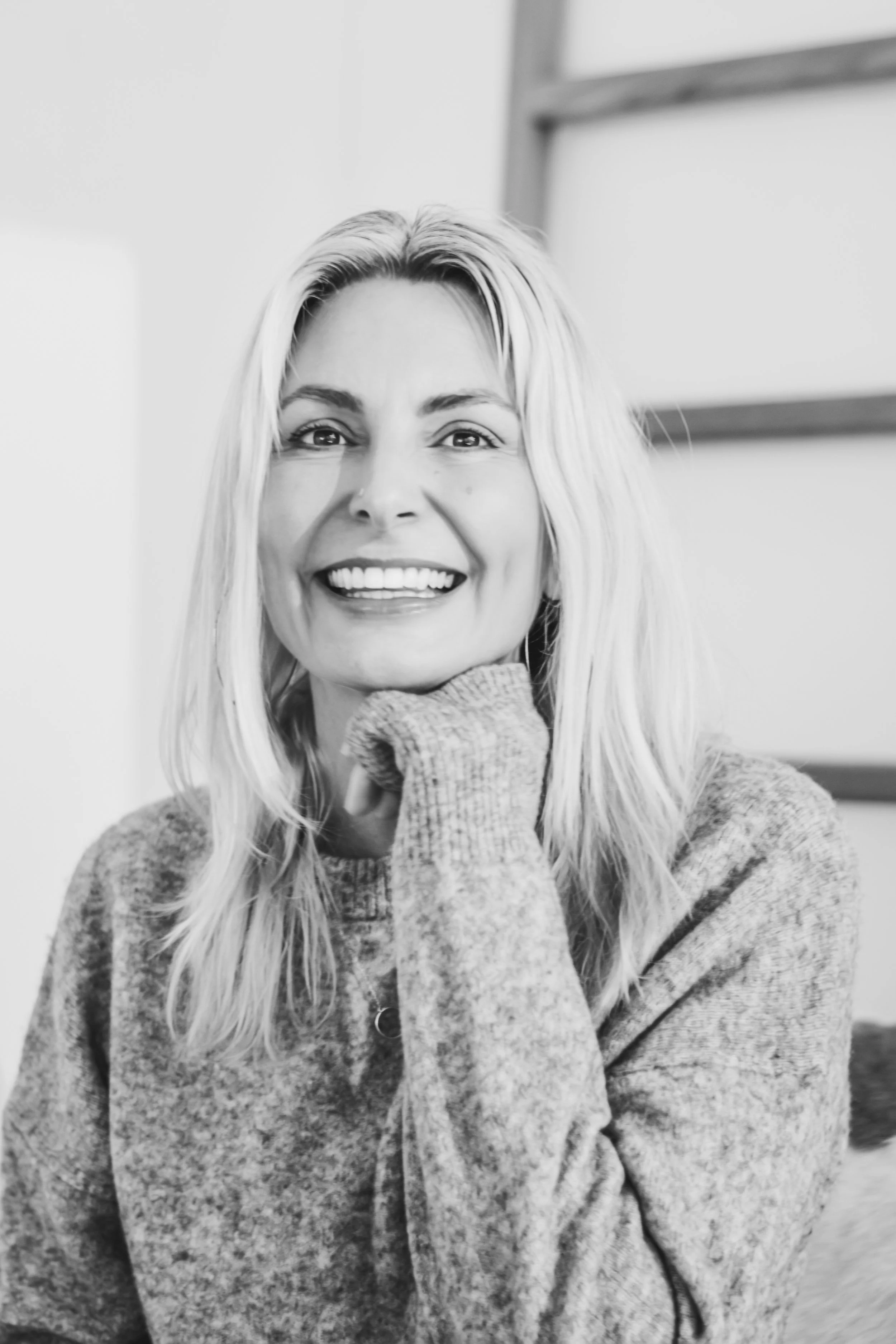 Black and white photo of a woman smiling with her hand resting under her chin, wearing a cozy sweater.