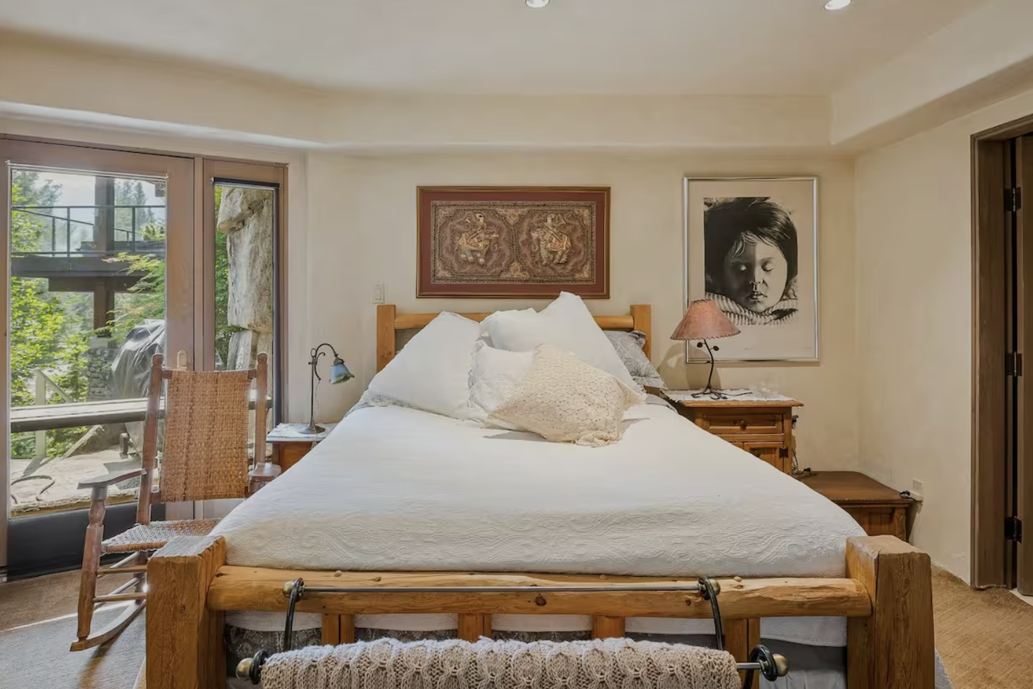 A bedroom with a large bed, wooden headboard, off-white bedding, and decorative pillows. There is a nightstand with a lamp and art on the wall, along with trees visible through sliding glass doors.