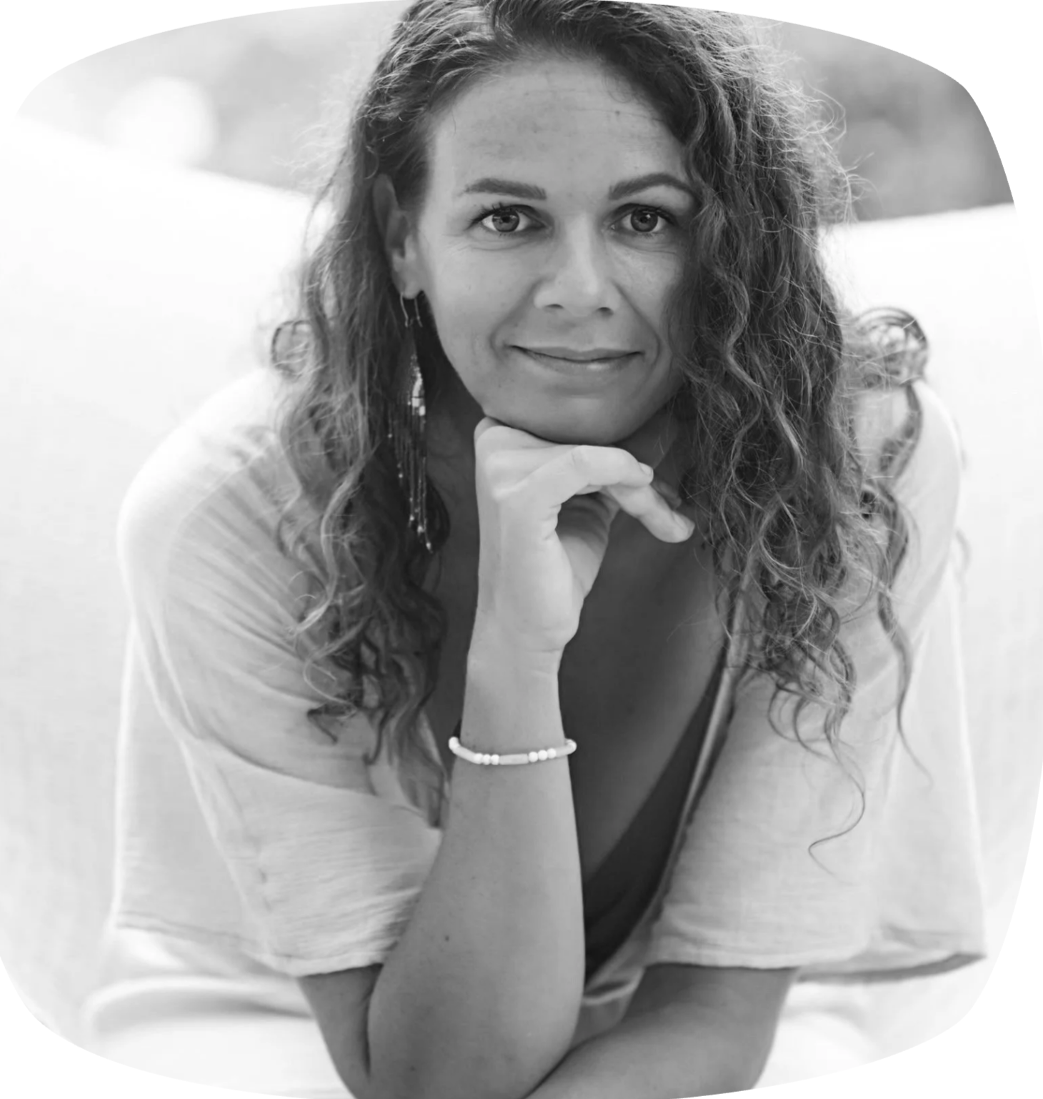 Black and white photo of a woman with curly hair, resting her chin on her hand, looking at the camera with a slight smile, wearing earrings and a bracelet.