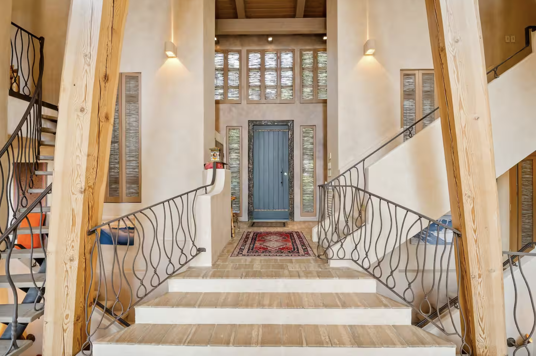 Entryway with staircase and wooden accents, featuring a blue door, decorative glass panels, a rug, and wall-mounted lights.