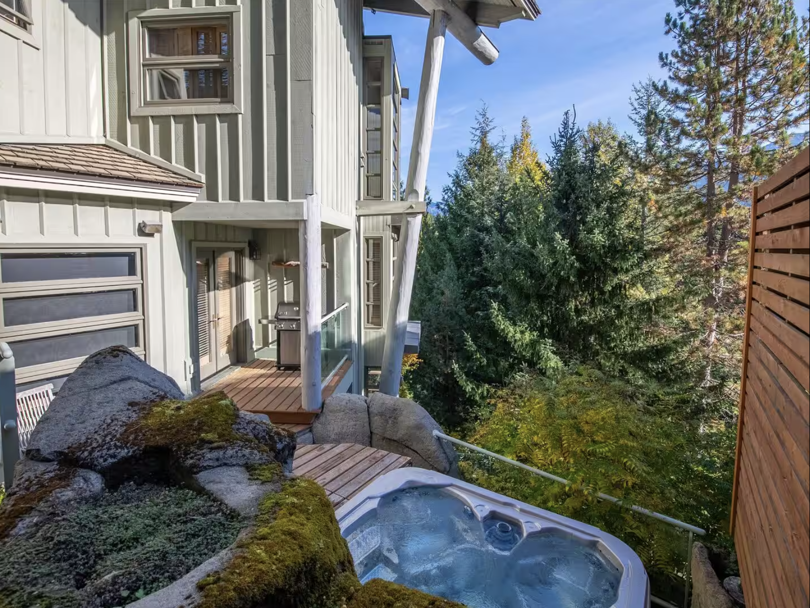 An outdoor deck adjacent to a house with large windows, a grill, a hot tub, and surrounded by green trees.