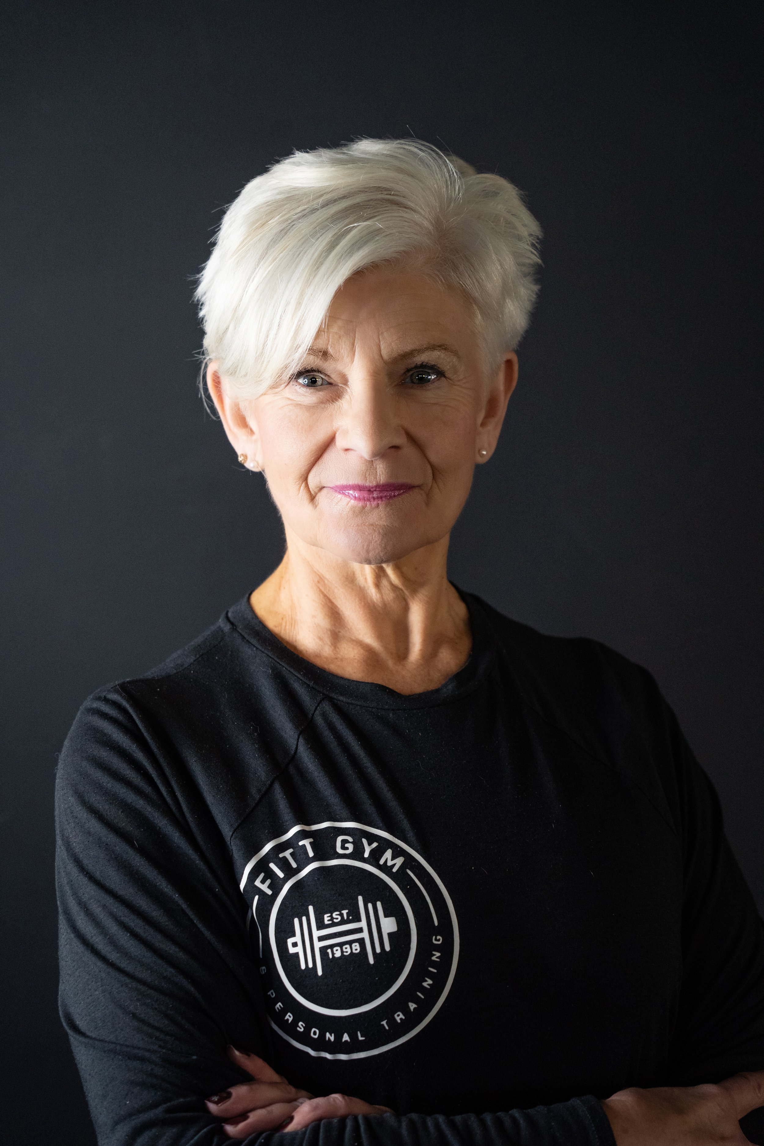 A confident elderly woman with short white hair, wearing a black fitness shirt with a logo, standing against a dark background.