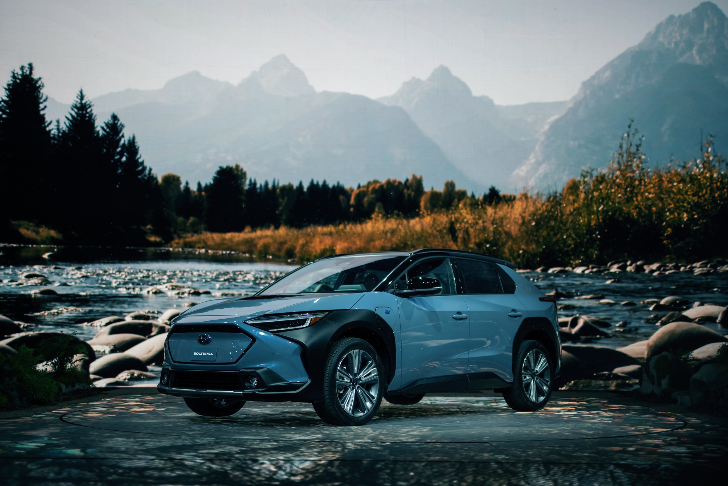 A blue electric car parked in front of a scenic mountain landscape with a river and trees.