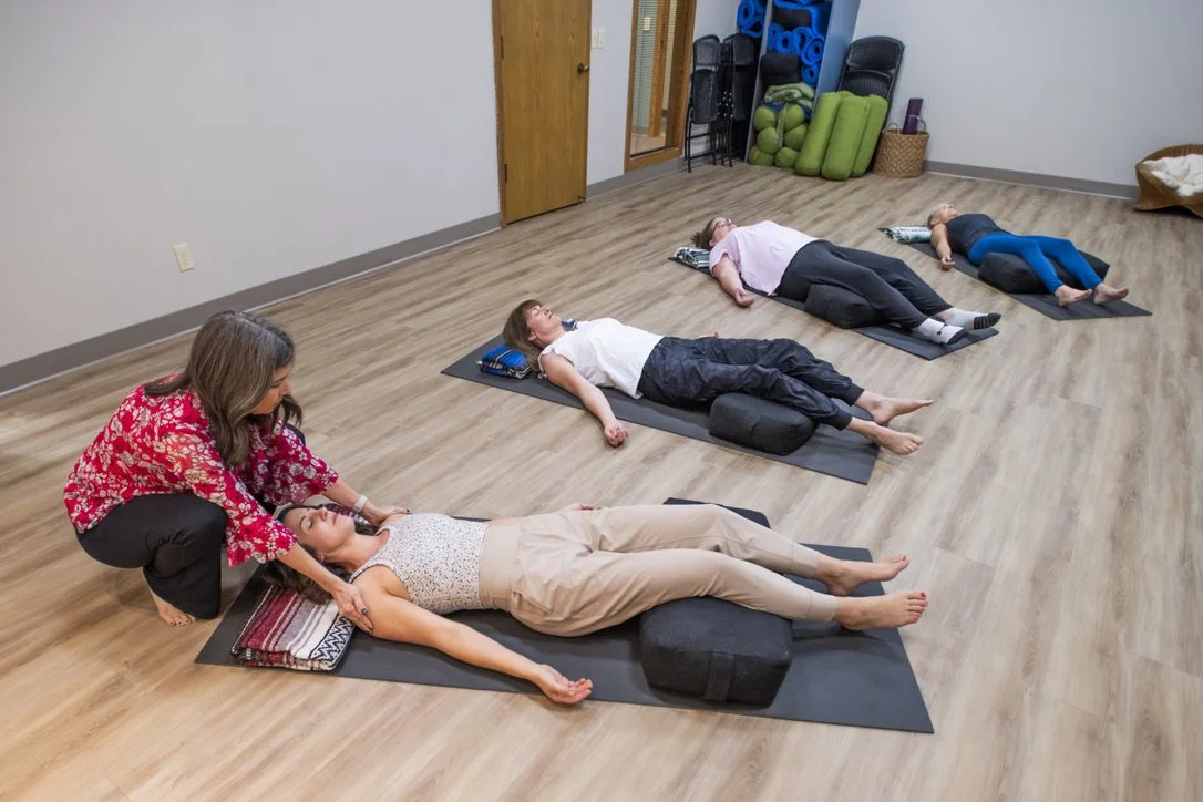 Instructor guiding a yoga class with four women lying on yoga mats in a spacious studio.