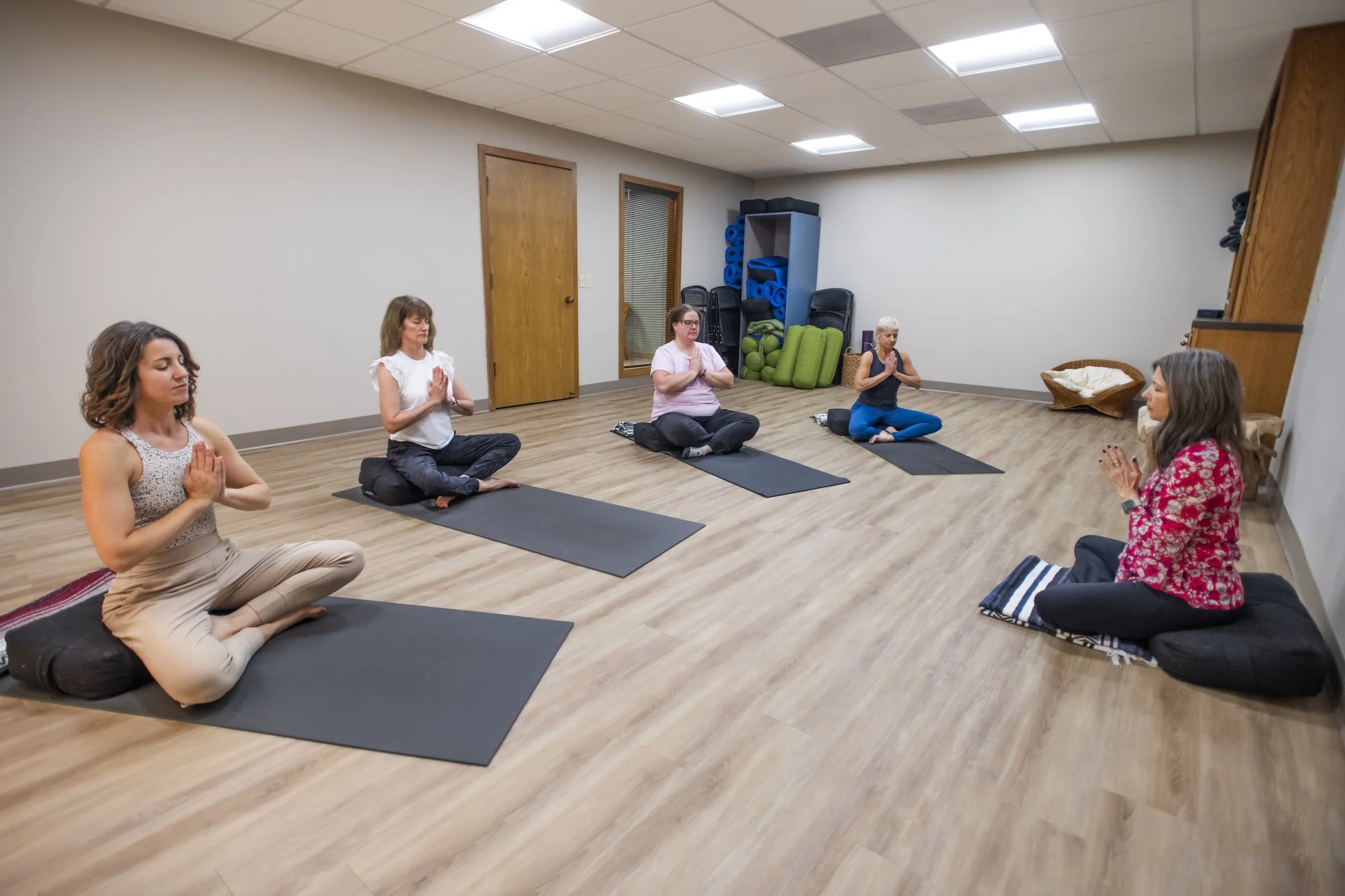 A group of five women participating in a seated meditation class in a room with wooden floors and white walls. They are sitting cross-legged on black mats with their hands in a prayer position.
