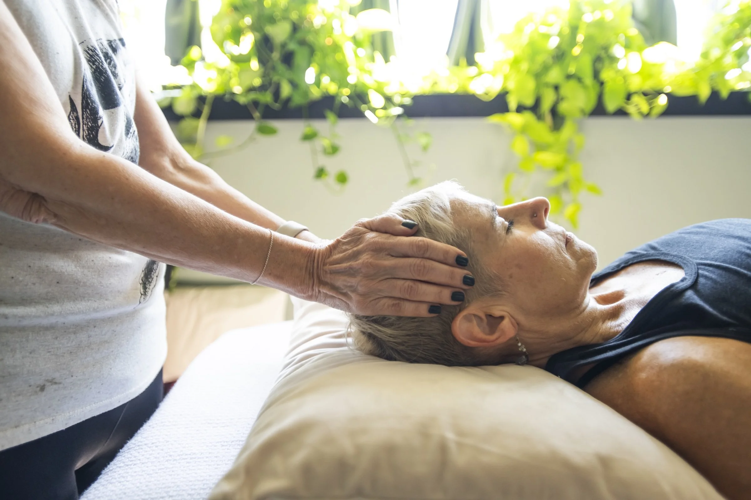 Person receiving a head massage from another woman in a space with natural light and green plants.