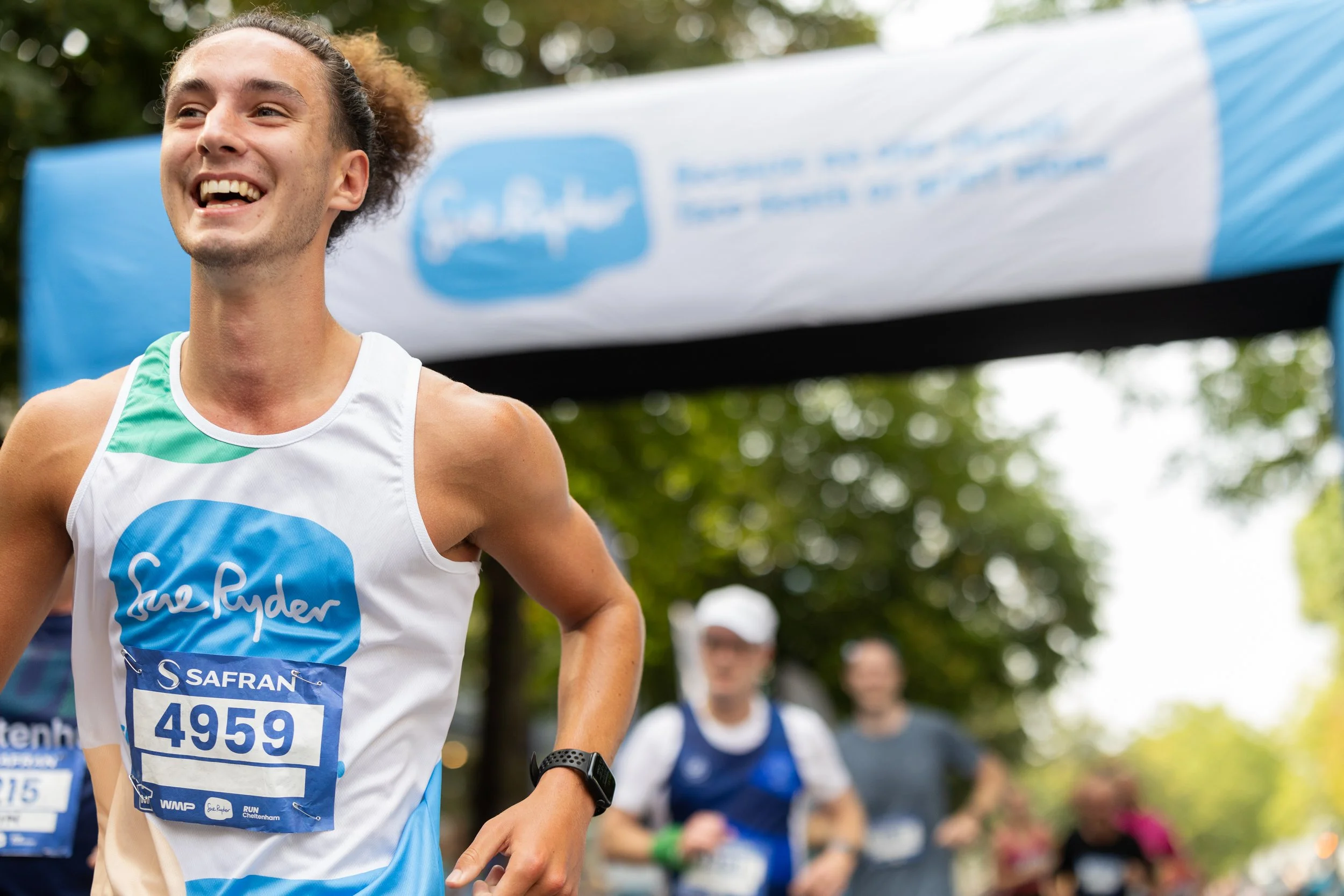 A young male runner smiling, wearing a white and blue tank top with a bib number 4959, finish line banner in the background, other runners behind him, trees, and a partly cloudy sky.
