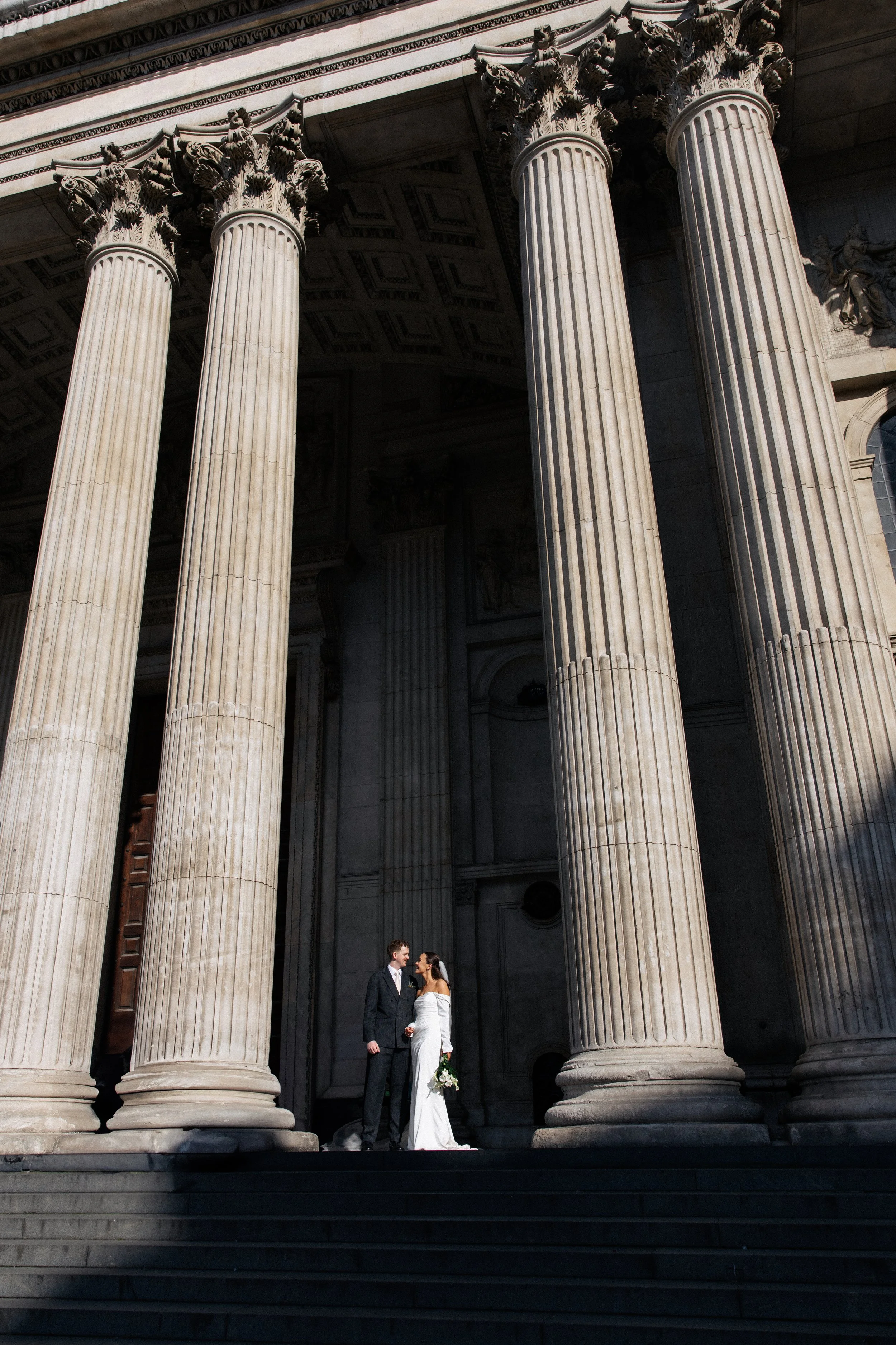 A bride and groom stand on the steps of St. Paul's Cathedral in the sunshine after their wedding