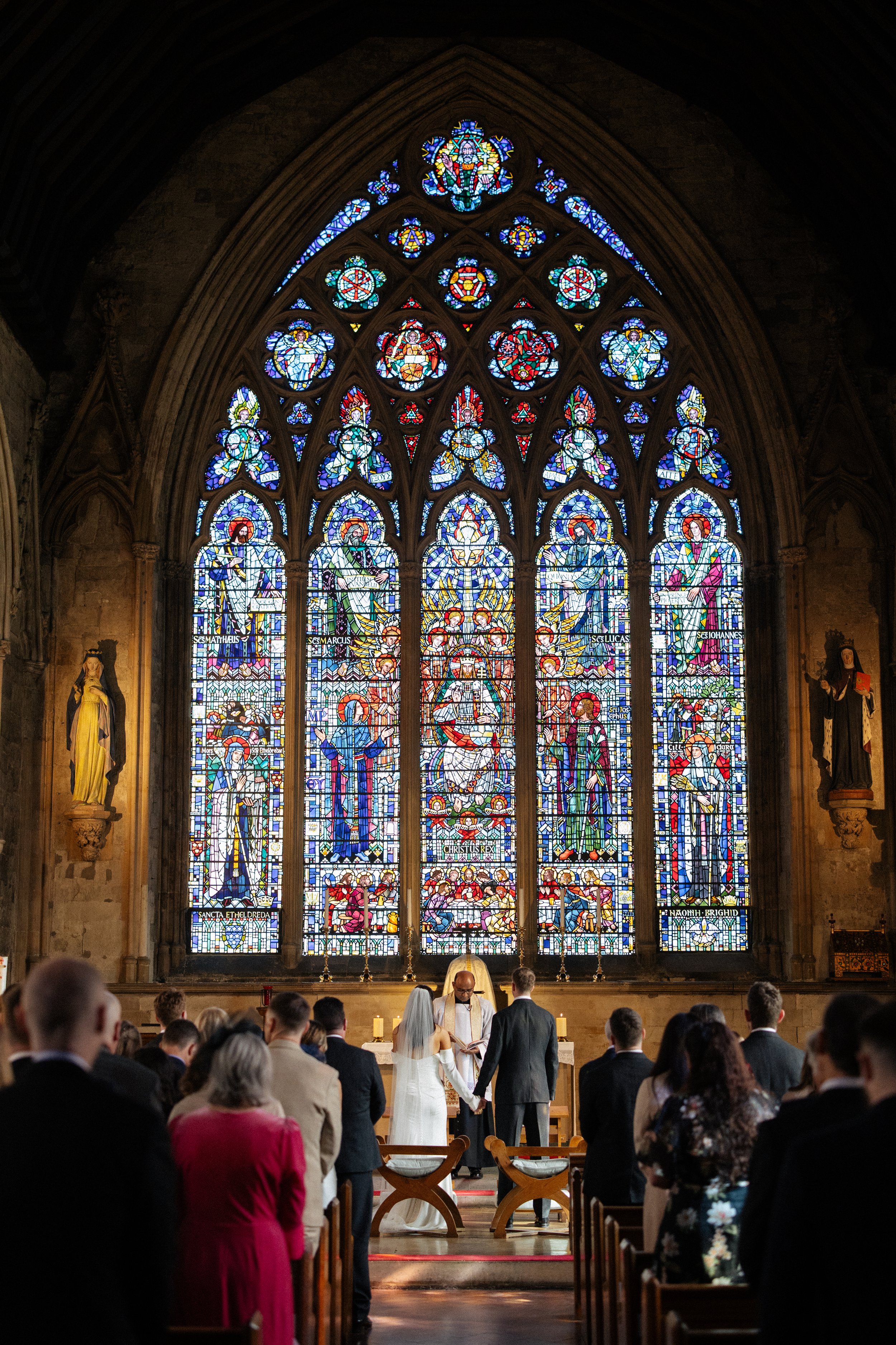 A couple stand in front of a stained glass window at St. Ethelreda's church during their wedding ceremony