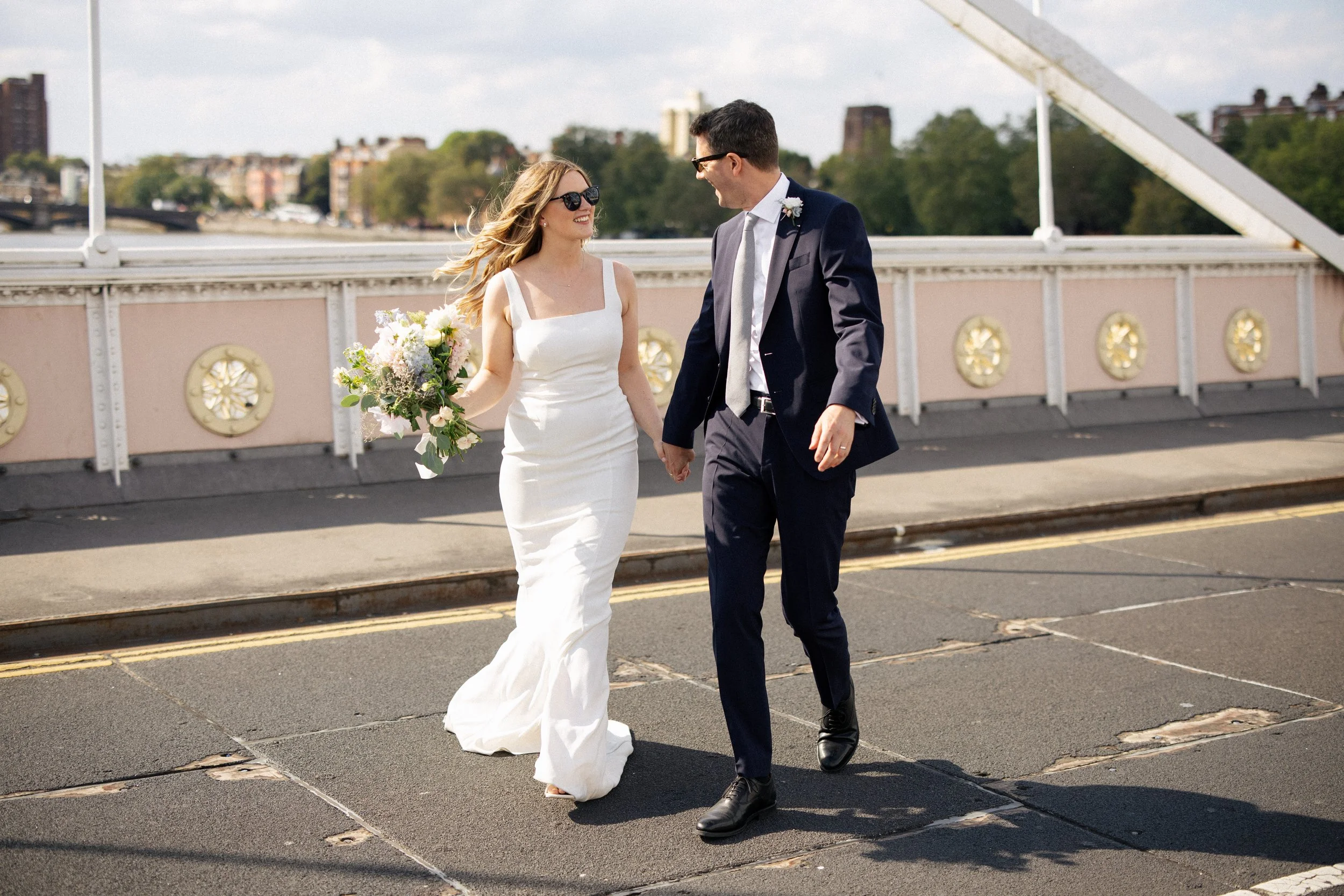 A bride and groom walk across the Albert Bridge together after their wedding at Battersea Park