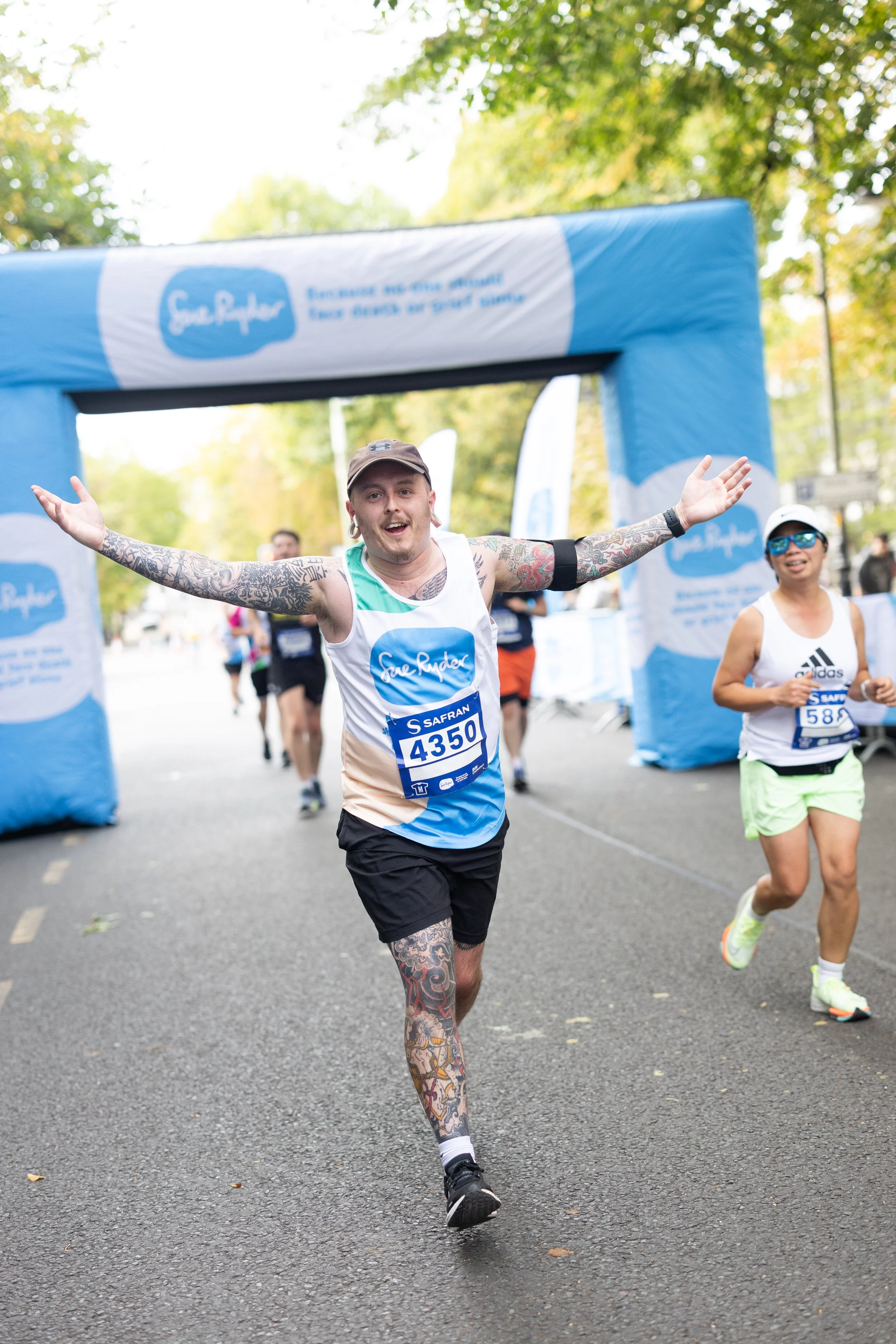A man with tattoos on his arms and legs, wearing a cap and a race bib number 4350, is running through a finish line at a race event, smiling and with arms outstretched. Other runners are visible behind him.