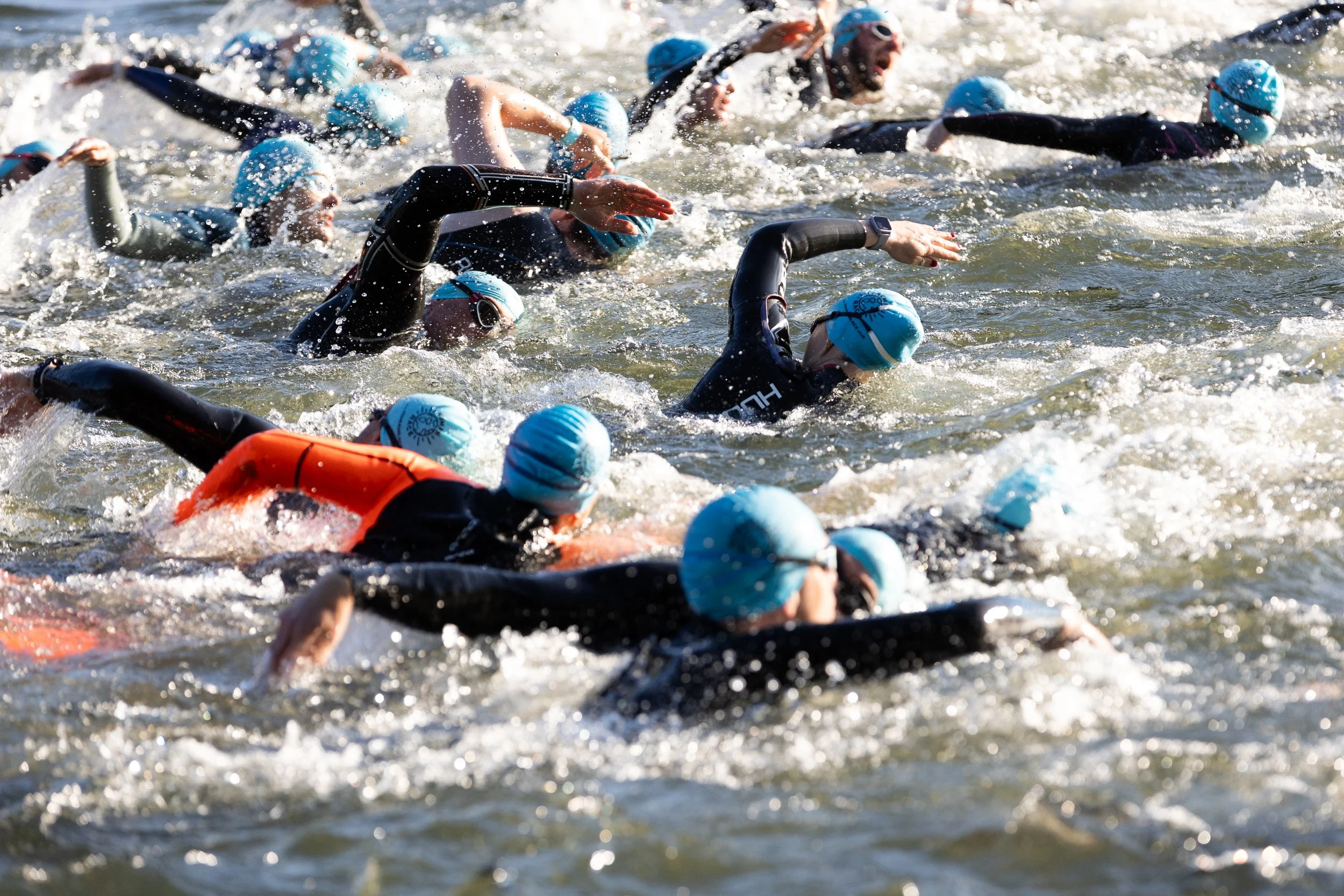 A group of swimmers, wearing black wetsuits and blue swim caps, competing in an open water swimming race, paddling through the water.