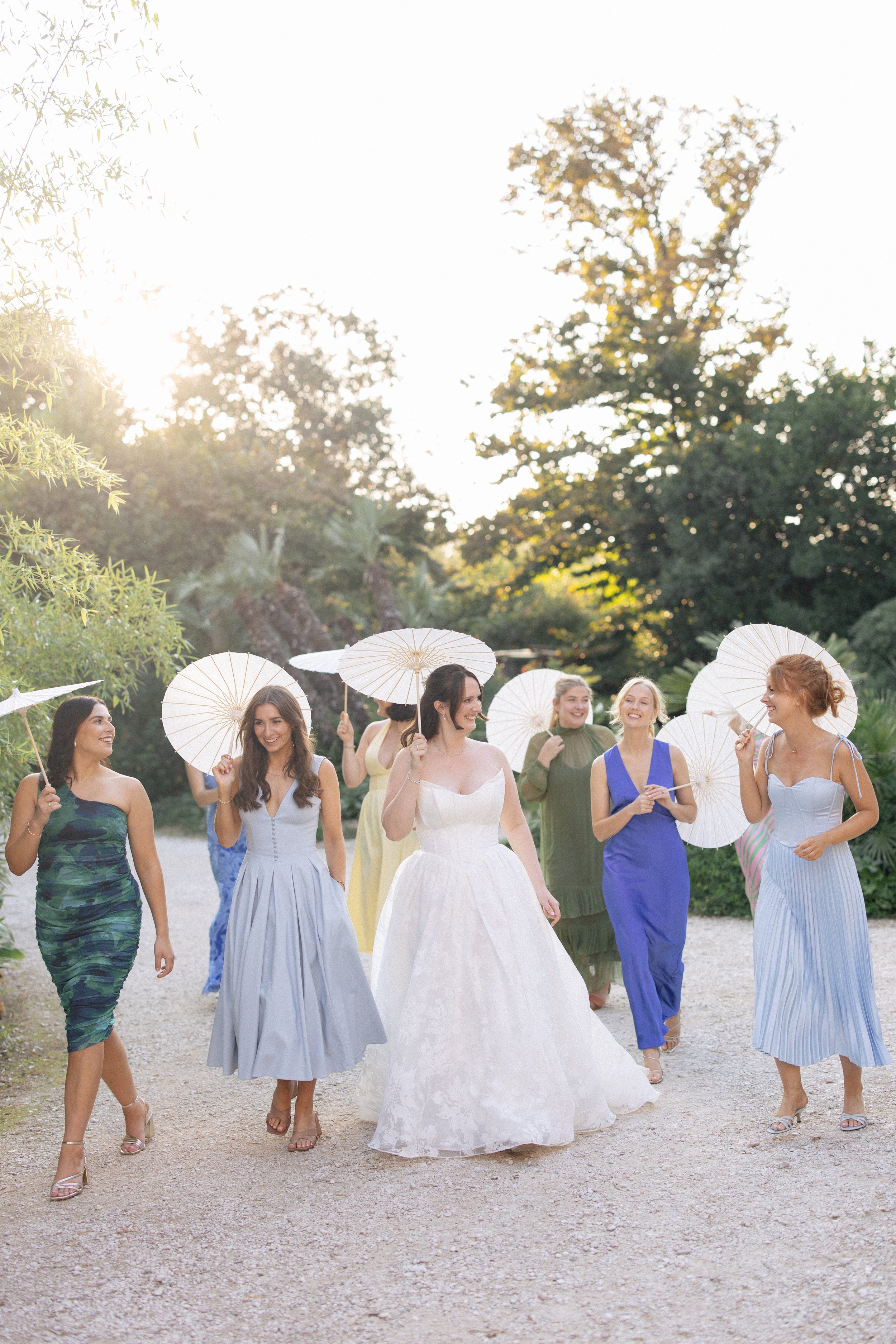 A bride and her friends walk through the sunset with parasols, laughing and joking at a wedding at Villa Giulia in Fano, Italy