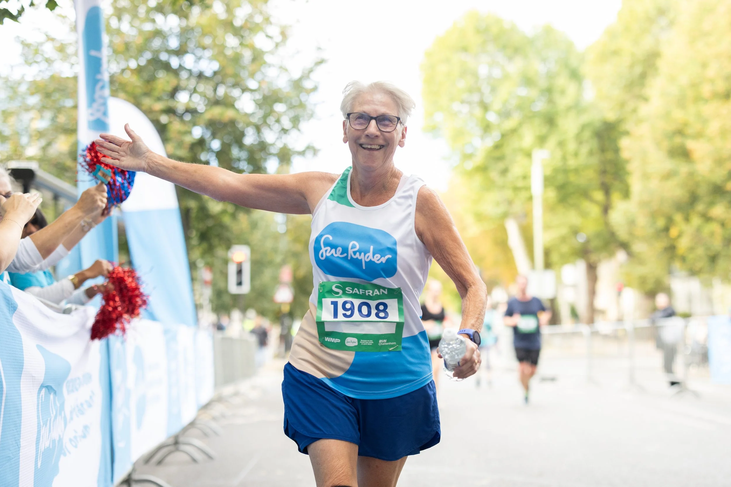 Older man running in a marathon, smiling, wearing glasses, a white tank top with a blue logo, blue shorts, and holding a water bottle. He is reaching out to high-five volunteers along the race route with a backdrop of trees and some other runners in 