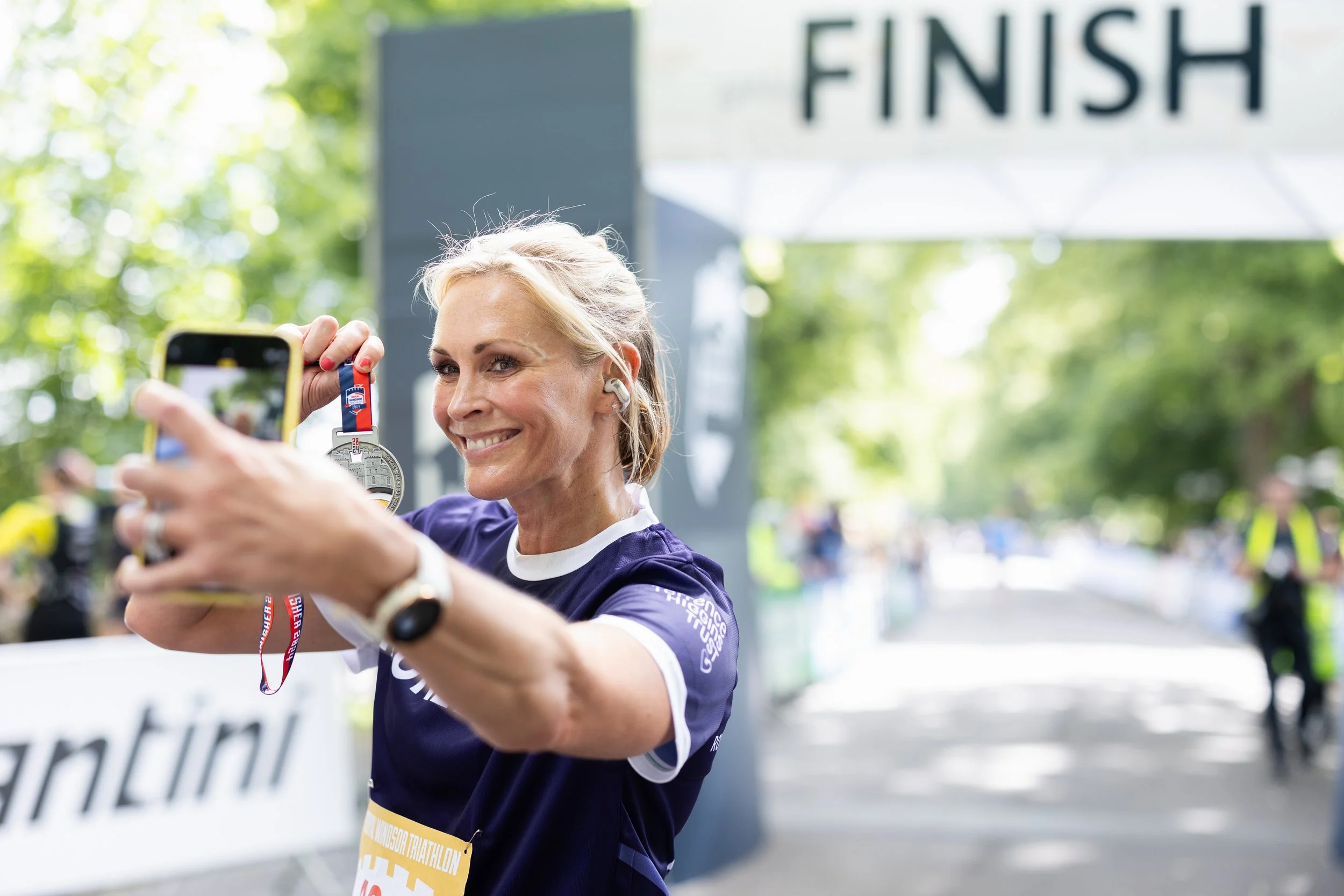 A woman at a race finish line taking a selfie while holding a medal, smiling, with a large 'FINISH' sign overhead, trees in the background, wearing a navy blue race shirt and earphones.
