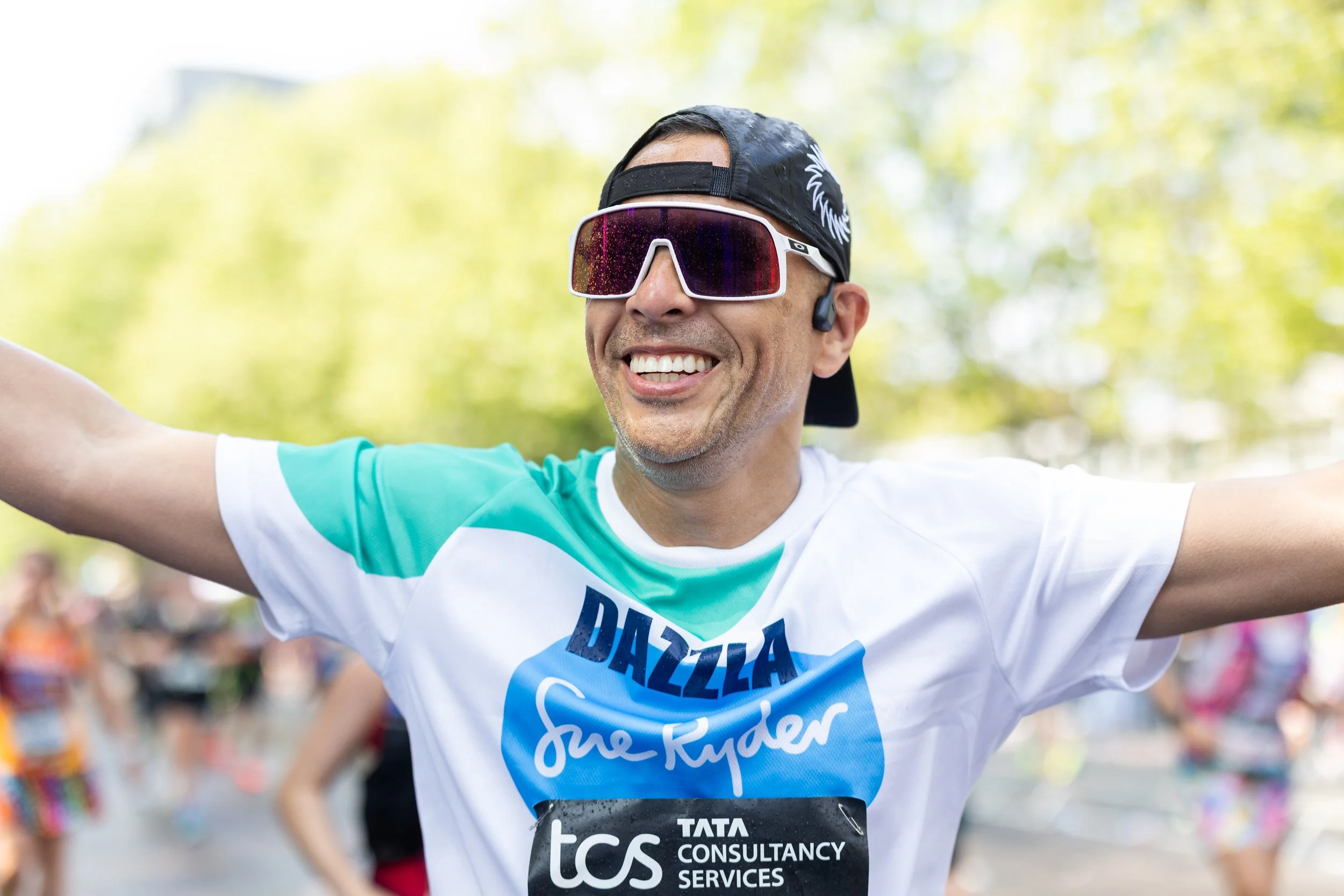 Smiling man wearing sunglasses, a black cap backwards, a teal and white T-shirt, and wireless earbuds, celebrating with his arms outstretched during a race or marathon outdoors.