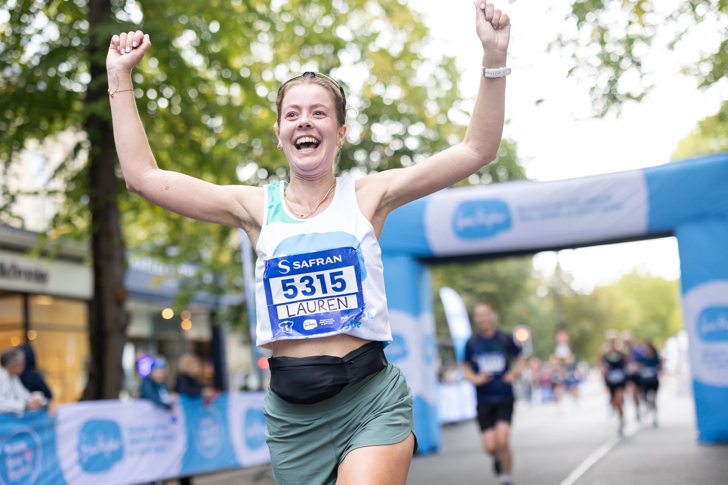 A woman with a race bib number 5315, named Lauren, celebrating as she crosses the finish line of a race with her arms raised in victory, surrounded by other runners and race banners, outdoors with green trees in the background.
