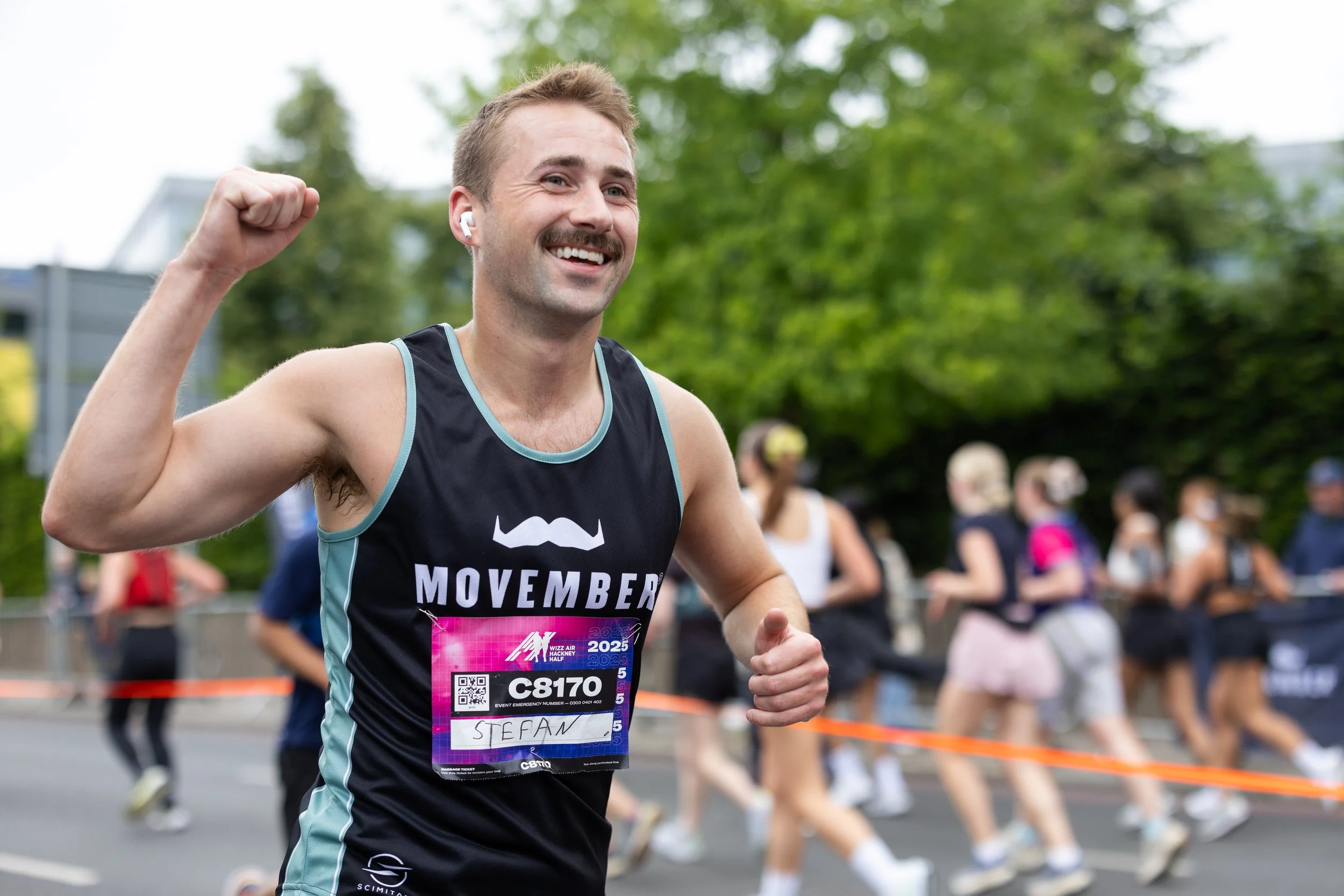 A man smiling and flexing his right arm while running in a marathon, wearing a black tank top with 'Movember' written on it, a race bib with the number C8170 and the name Stefan, with other runners and green trees in the background.
