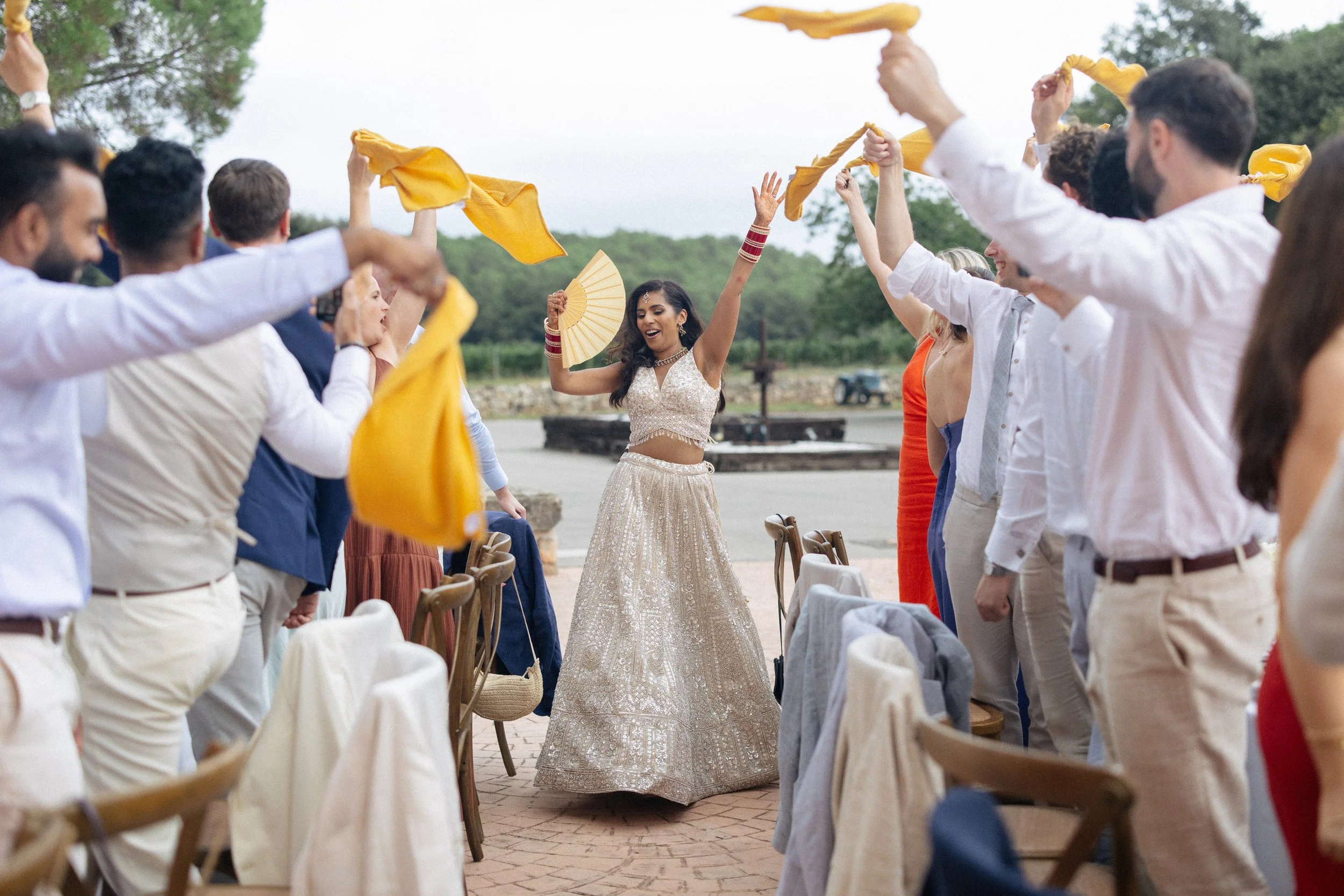 A bride in traditional Indian wedding dress walks down between tables of wedding guests swinging their napkins and cheering at Mas Del Sord, Spain