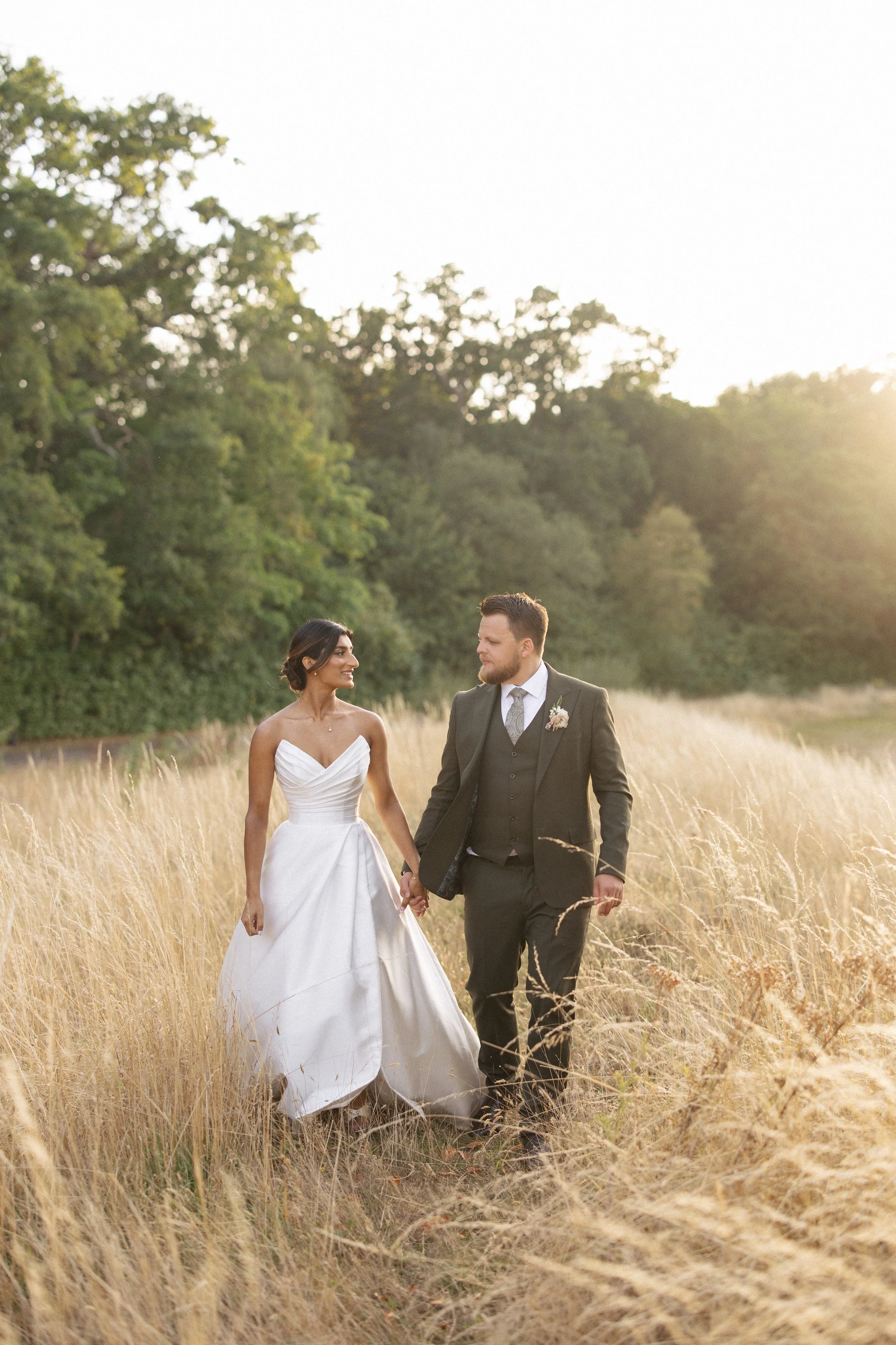 A bride and groom walk through long, golden grass at sunset after their wedding at Botleys Mansion
