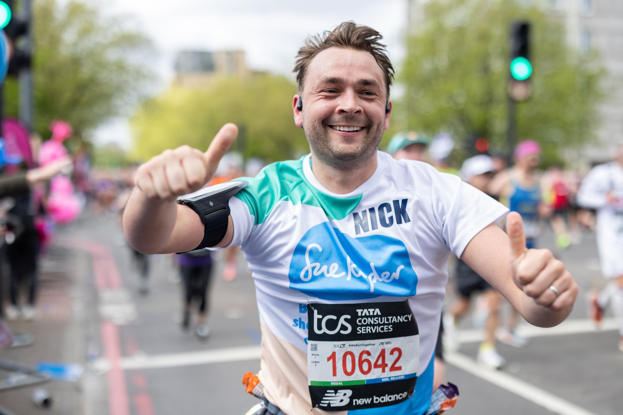 Man smiling with thumbs up at a marathon, wearing race bib number 10642, with fellow runners and trees in the background.