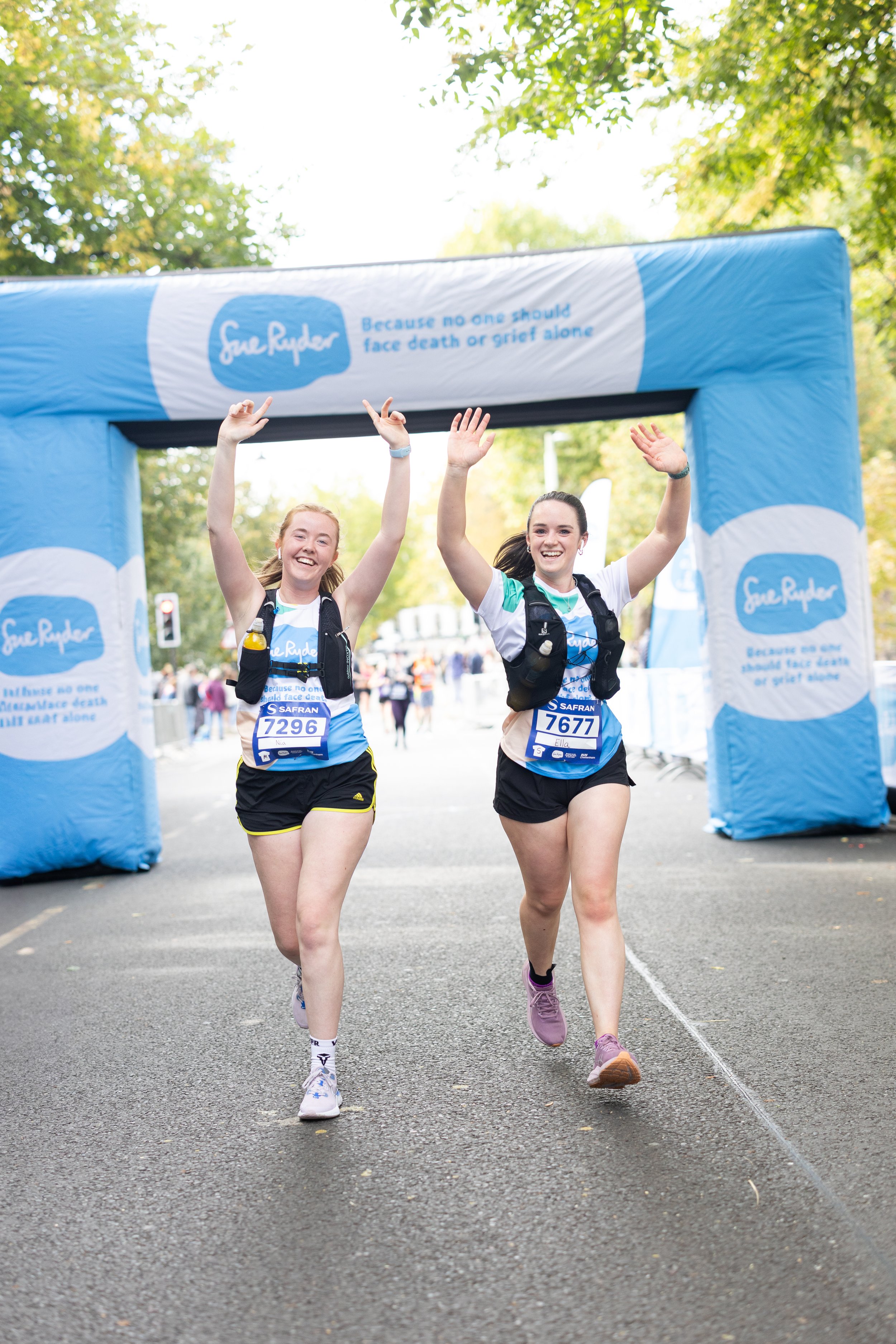 Two women participating in a marathon finish line event, crossing the finish line with arms raised and smiling, under an inflatable arch with blue and white branding.