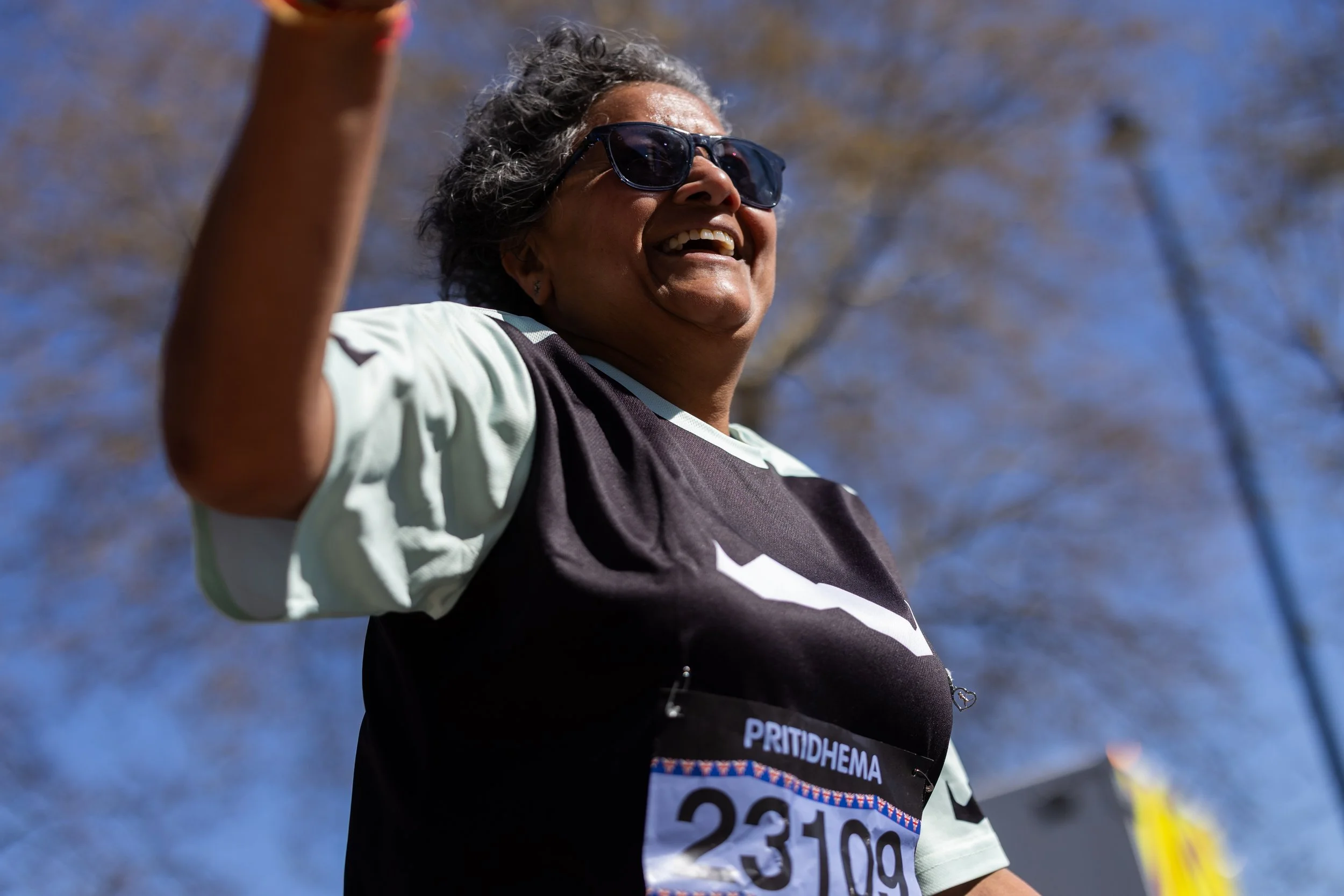 A smiling woman wearing sunglasses and a race bib, waving outdoors on a sunny day with trees in the background.
