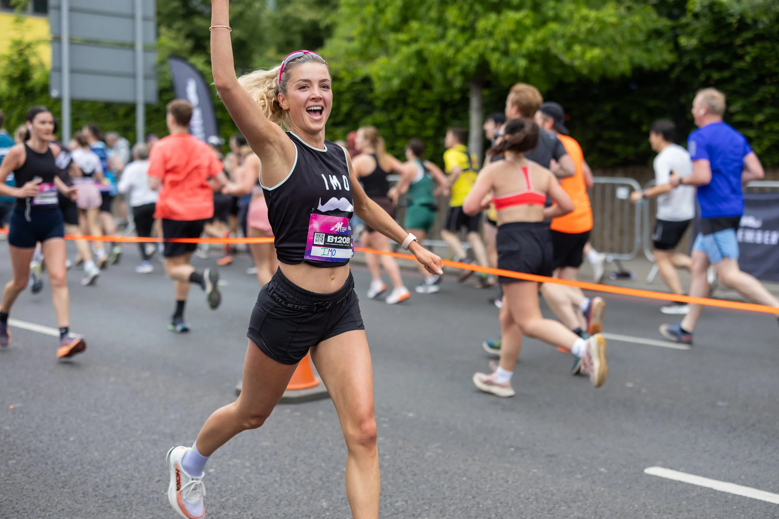 A young woman with blonde hair wearing a black running outfit and pink headband, smiling and crossing the finish line in a race, with other runners in the background.