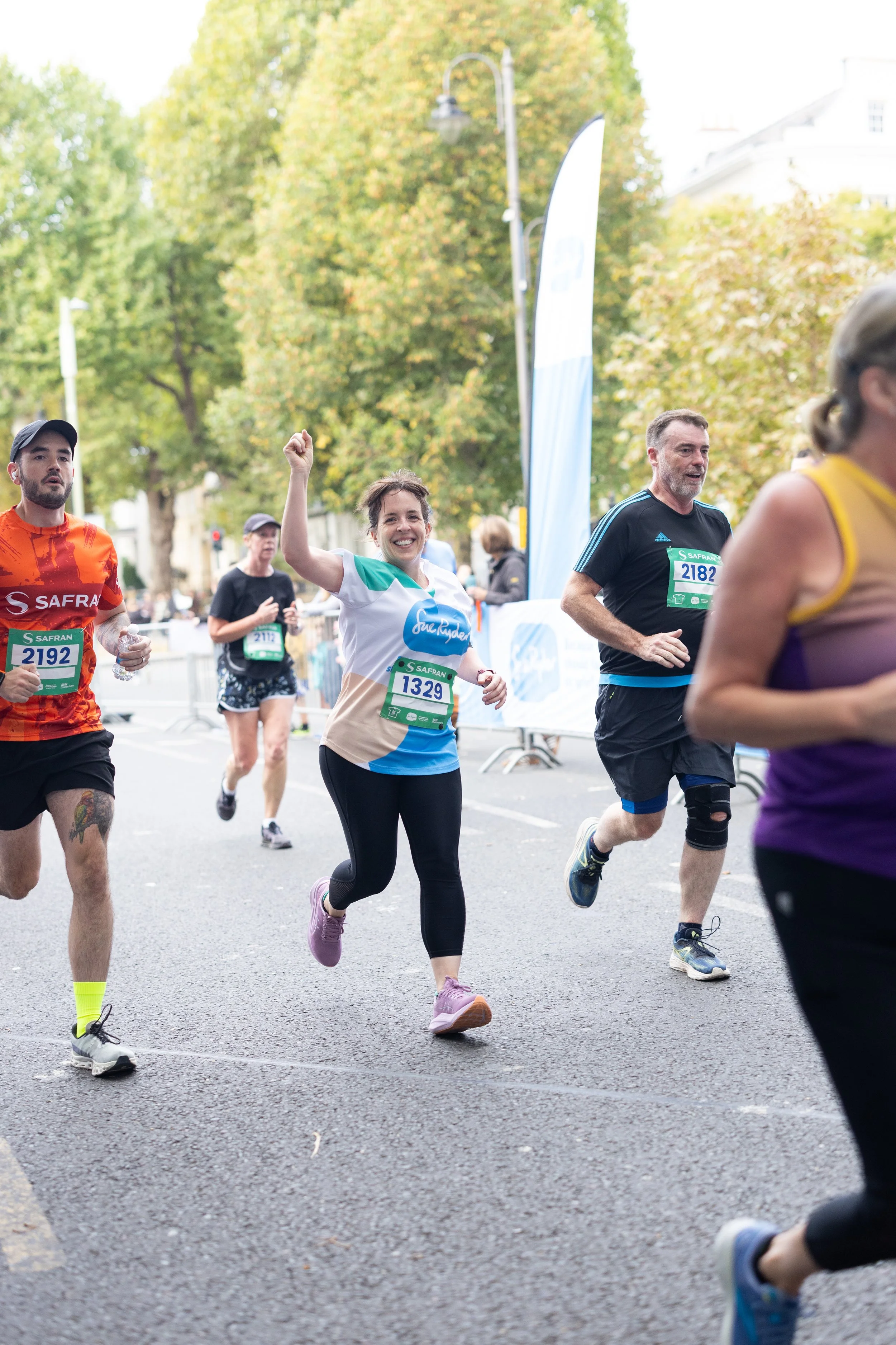 People running in a marathon, smiling and appearing energetic, with trees and race banners in the background.