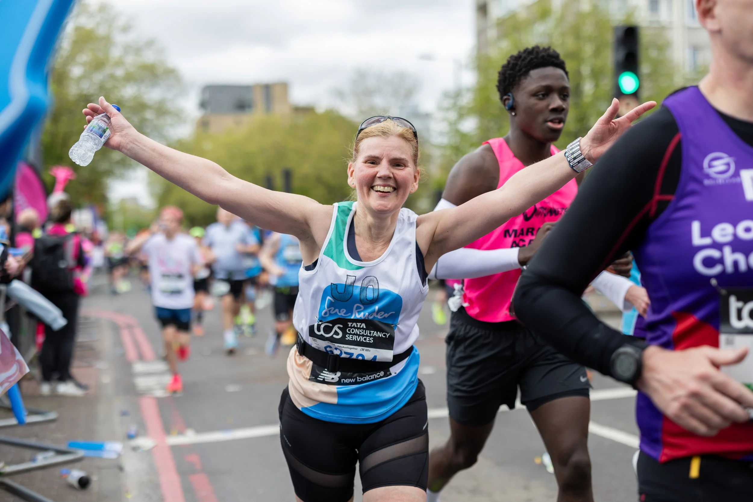 A smiling woman with blonde hair, wearing a white and blue race shirt, is running in a marathon with her arms outstretched. Several other runners are visible in the background on a city street.