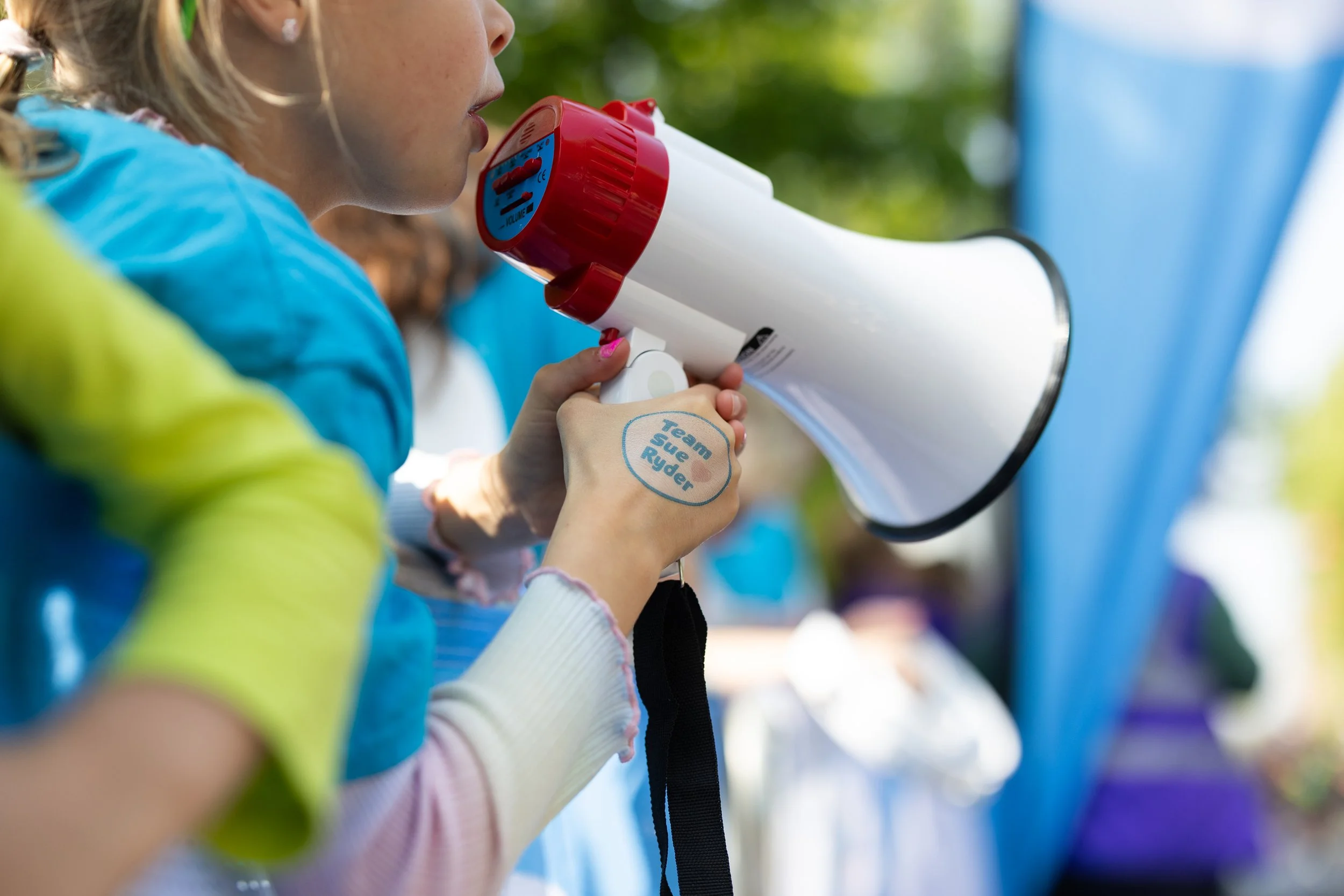 A young girl in a blue jacket speaking into a megaphone during an outdoor event, with other people blurred in the background.
