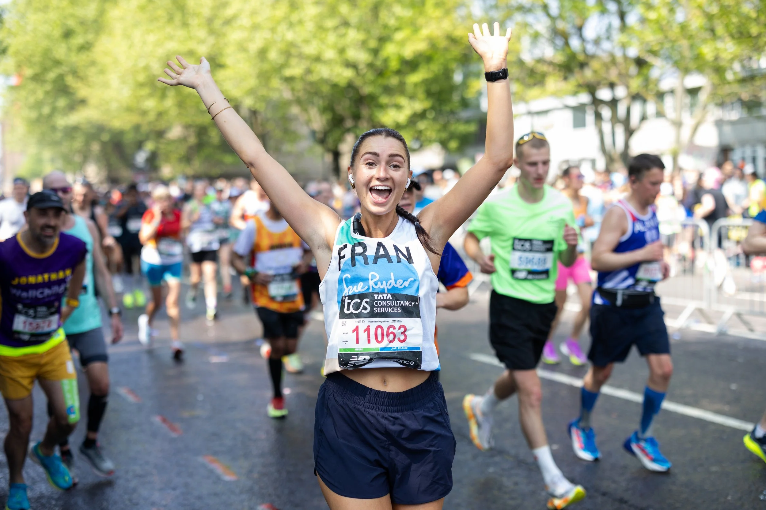 A young woman running in a marathon with her arms raised, smiling, surrounded by other runners on a city street with trees and buildings in the background.