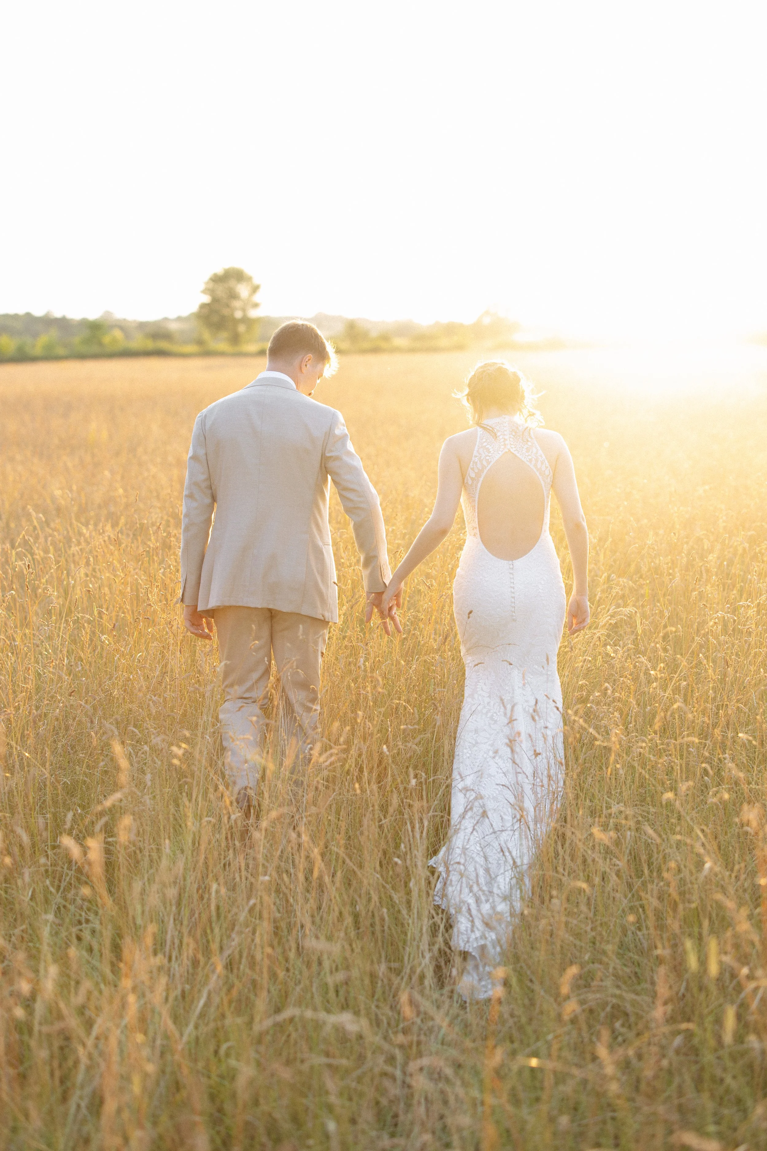 A bride and groom walk through long grass at sunset at Alpheton Hall Barns