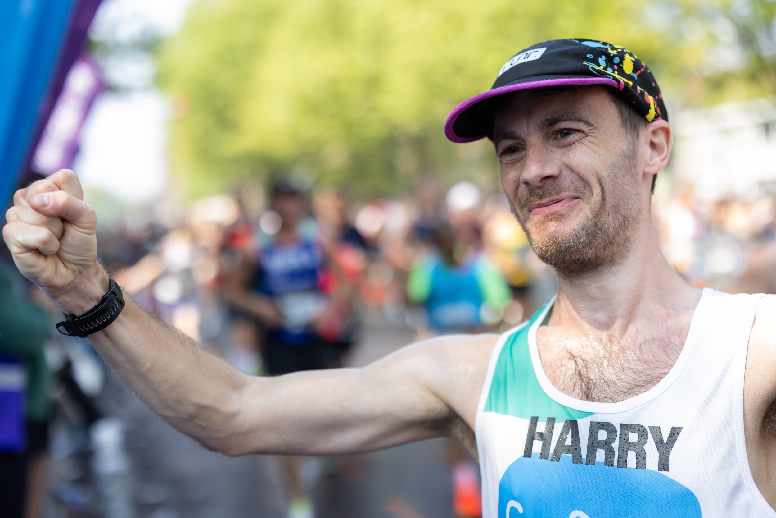 Man in athletic tank top and colorful cap celebrating after a race with a smile, raised arm, and clenched fist, with a blurred background of other runners and trees.