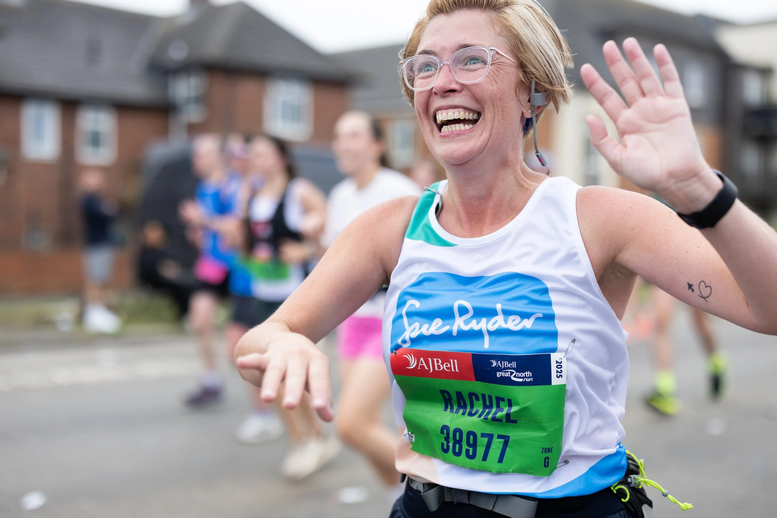 A cheerful woman smiling and waving during a running race, wearing a race bib with the name 'Rachel' and the number 38977, outside in an urban neighborhood with other runners in the background.