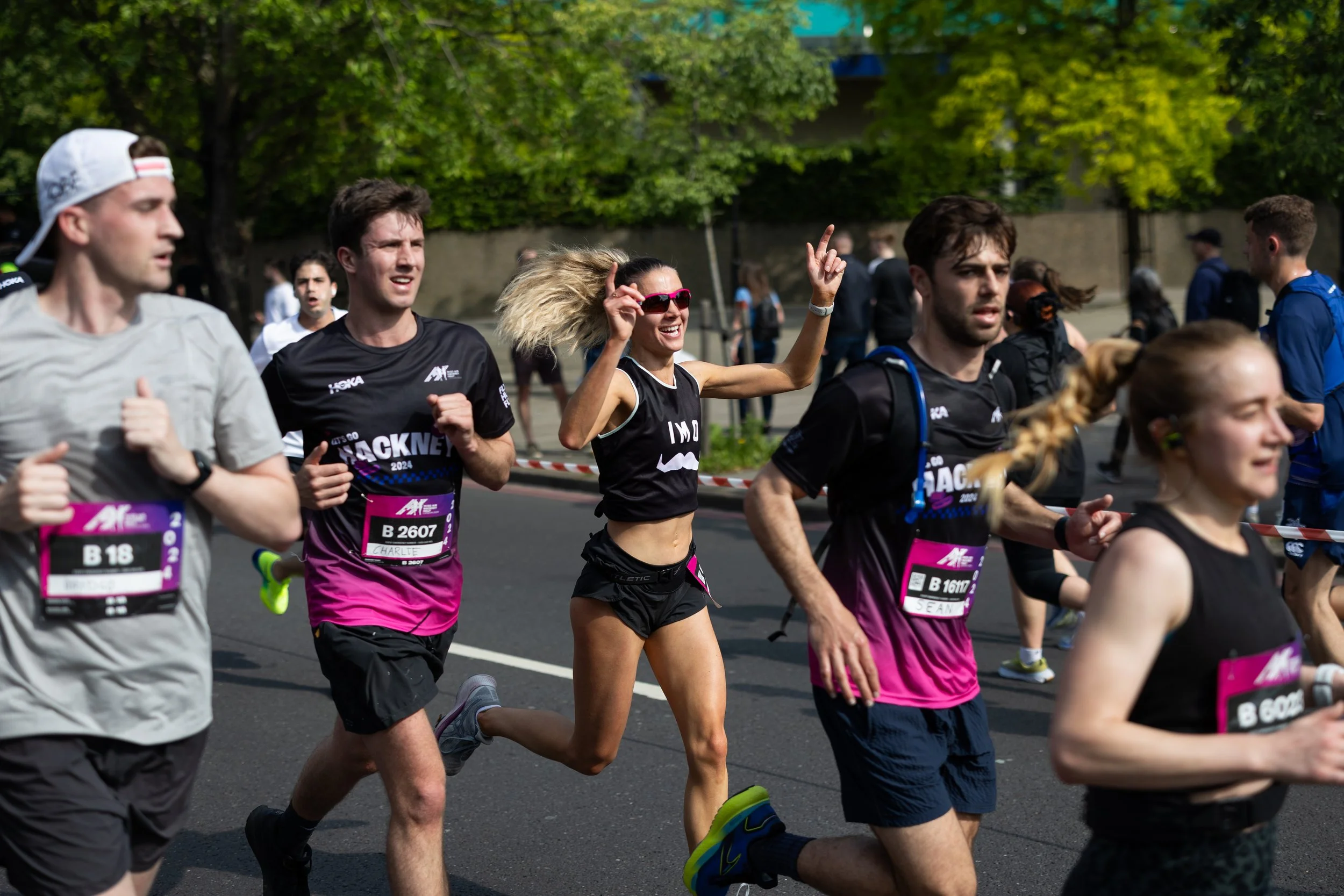 Group of runners participating in a marathon on a city street, wearing athletic clothing and race bibs, with trees and spectators in the background.
