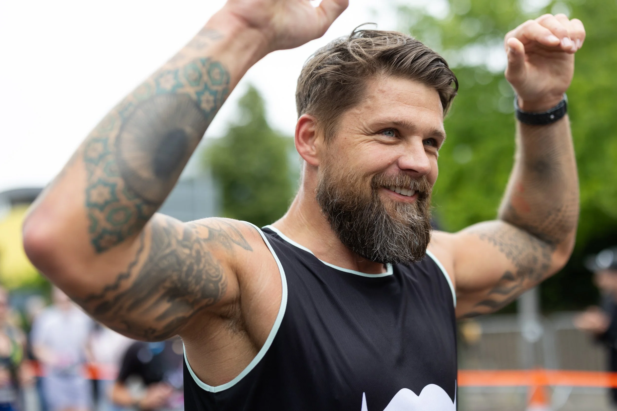 A man with a beard and tattoos smiling and flexing his biceps outdoors, wearing a black sleeveless shirt.