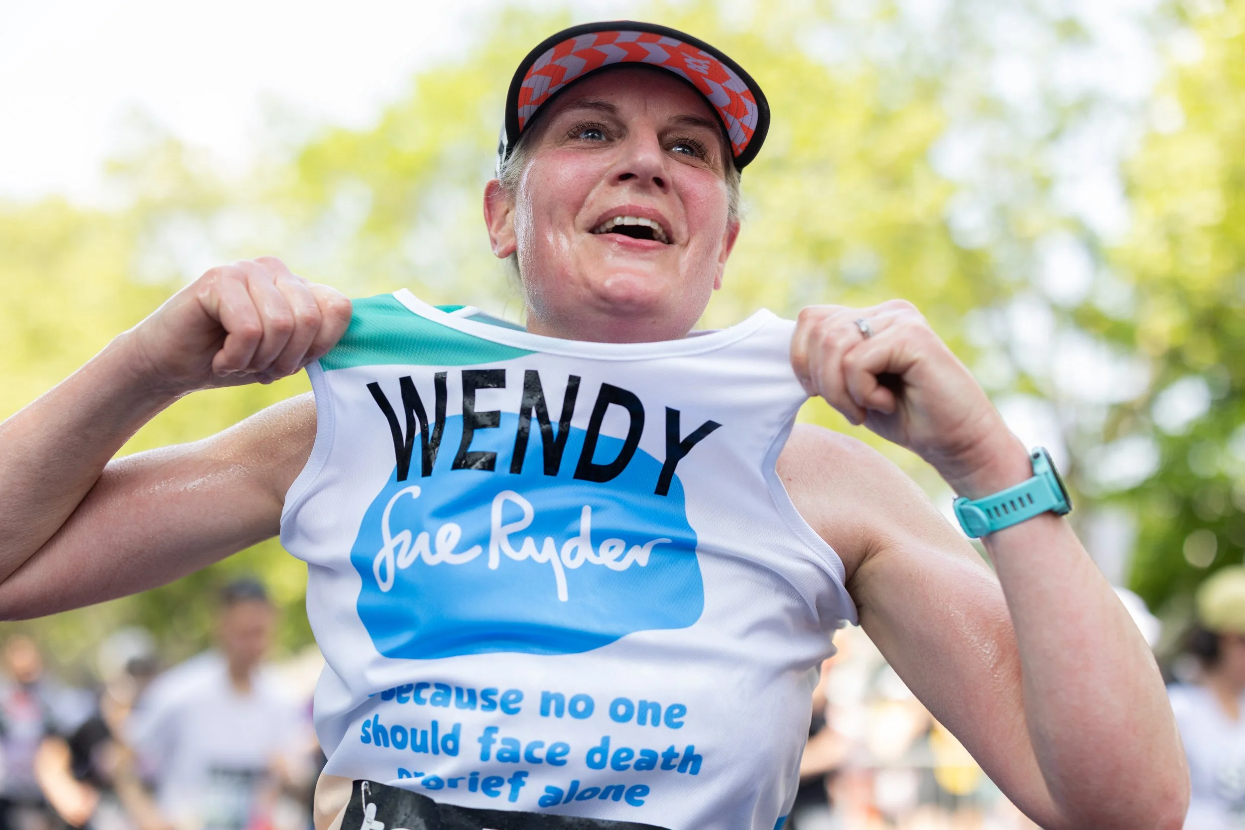 A woman wearing a running cap and a white race shirt with blue and black text, holding up her shirt with her fists during a marathon or race event. The background shows trees and other runners.