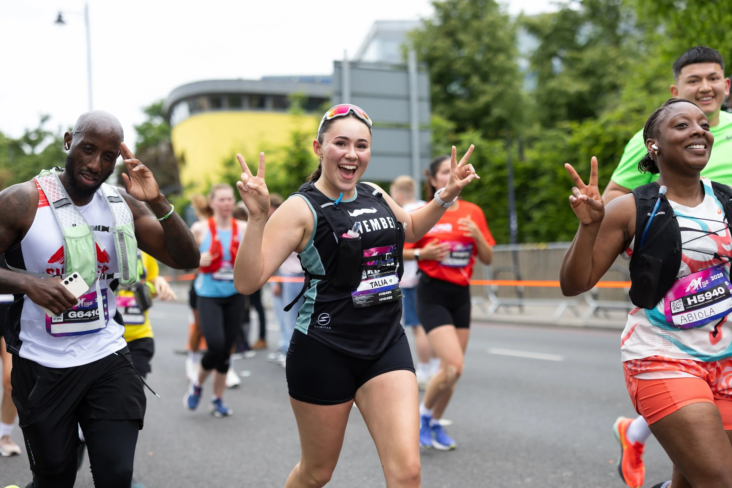 A group of marathon runners on a street during a race, smiling and making victory signs. Participants are wearing workout gear and bibs, with some holding water bottles.