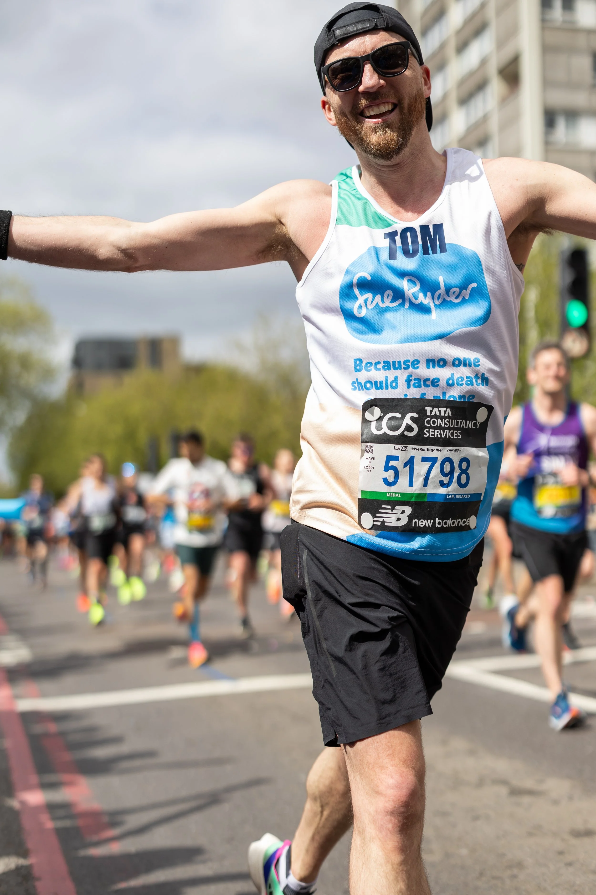 A man in a tank top, shorts, sunglasses, and a backward cap running in a marathon. He is smiling with his arms outstretched, surrounded by other runners.